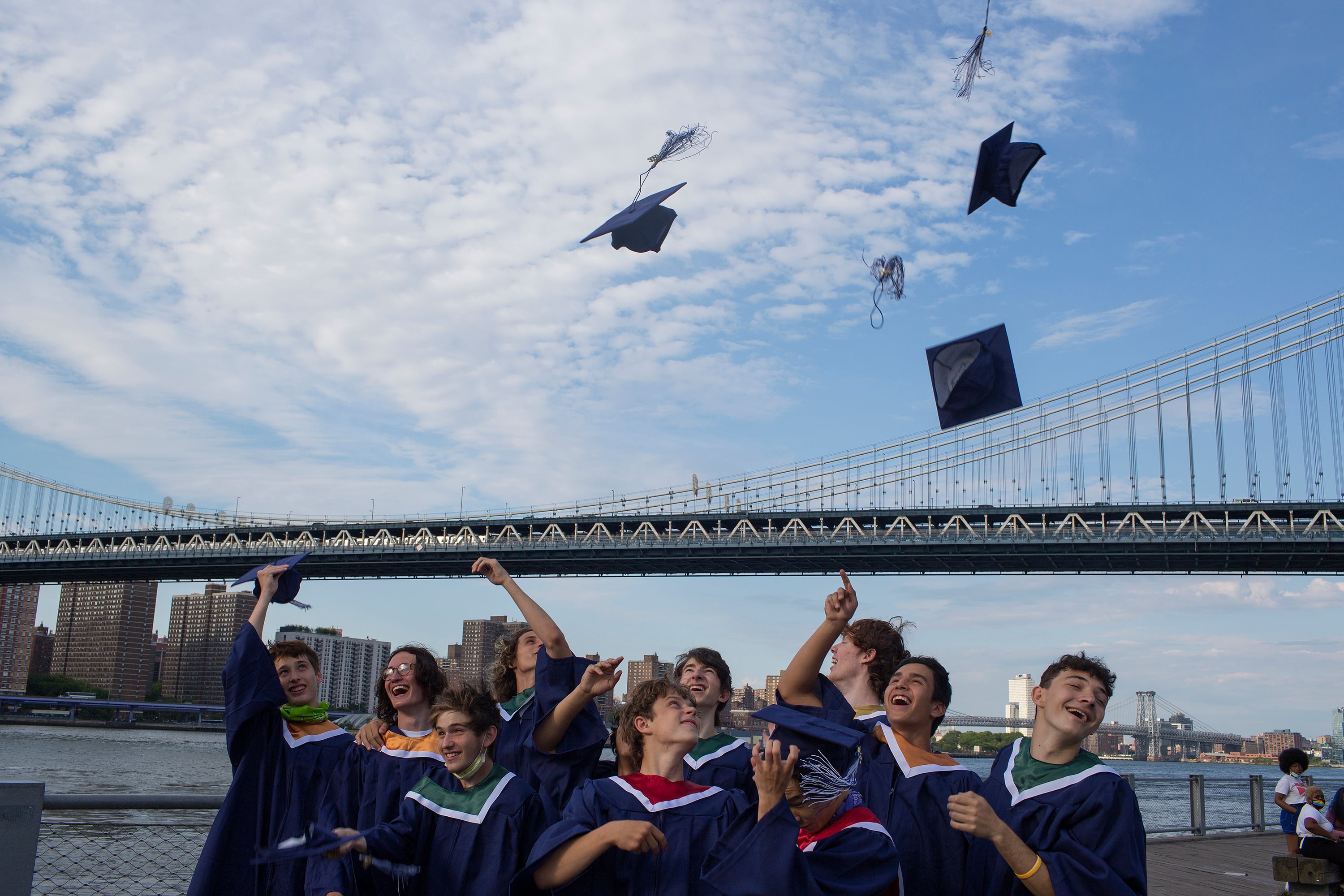 A group of high schoolers throw their graduation caps up in the air with the Brooklyn Bridge and a blue sky in the background.