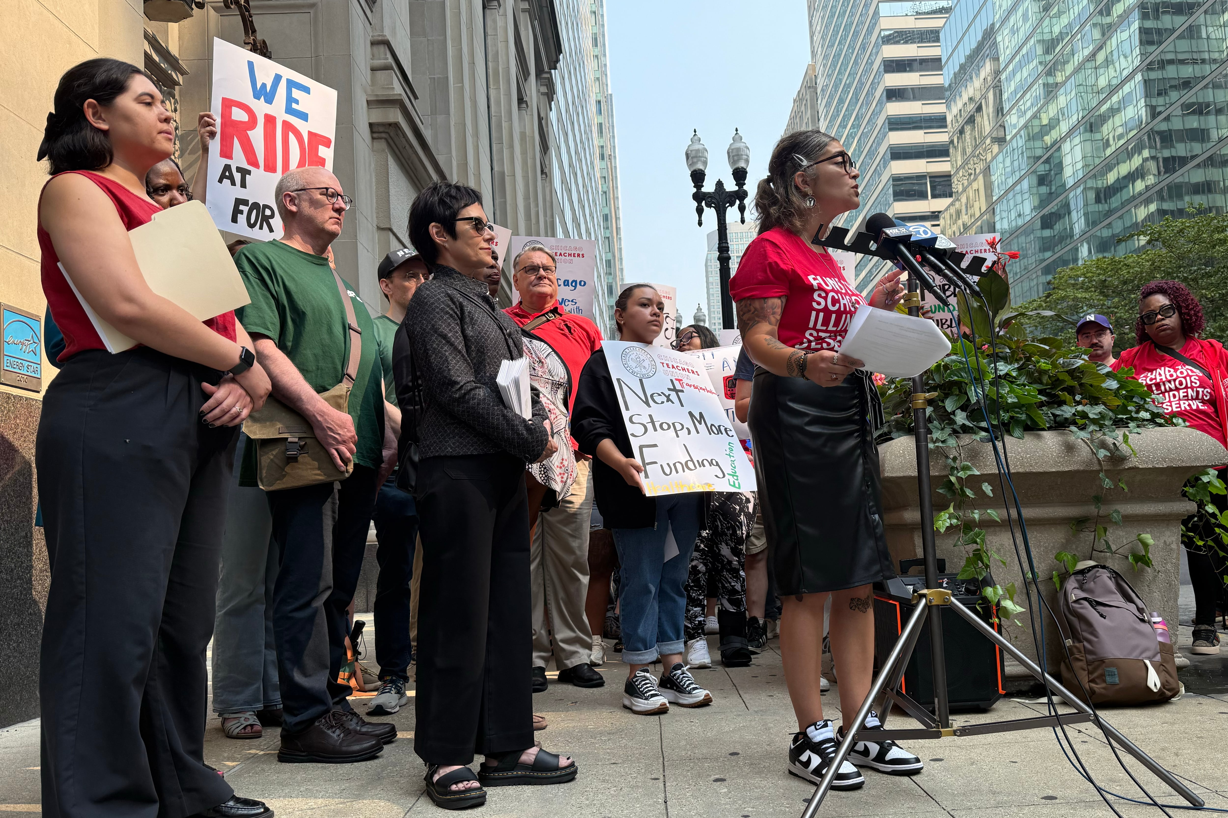 A photograph of a large group of people standing outside, some holding signs, while one person speaks from a microphone.