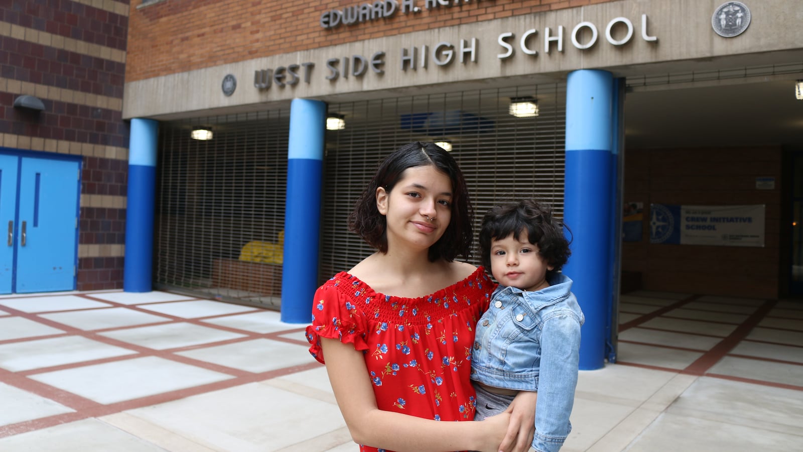 A woman in a red shirt holds a small child in a jean jacket in front of a brick school building with blue pillars.