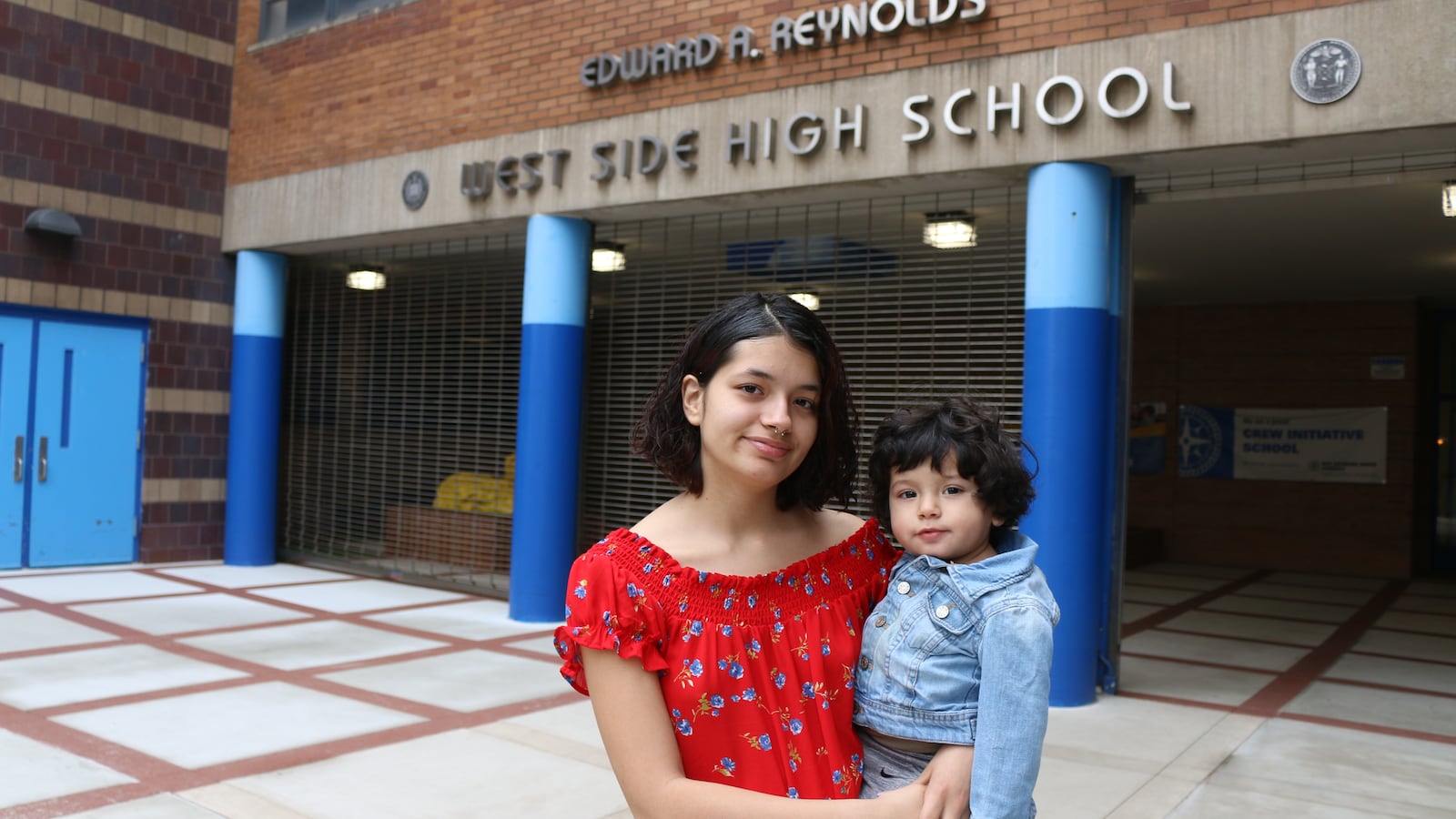 A woman in a red shirt holds a small child in a jean jacket in front of a brick school building with blue pillars. 