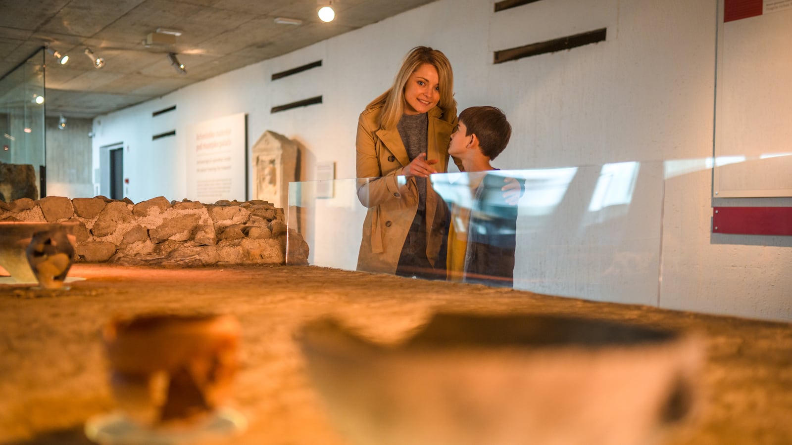 Woman and her son stand behind a glass partition and looking at a replica of a Roman restaurant. Waist up image, partly shot through glass.