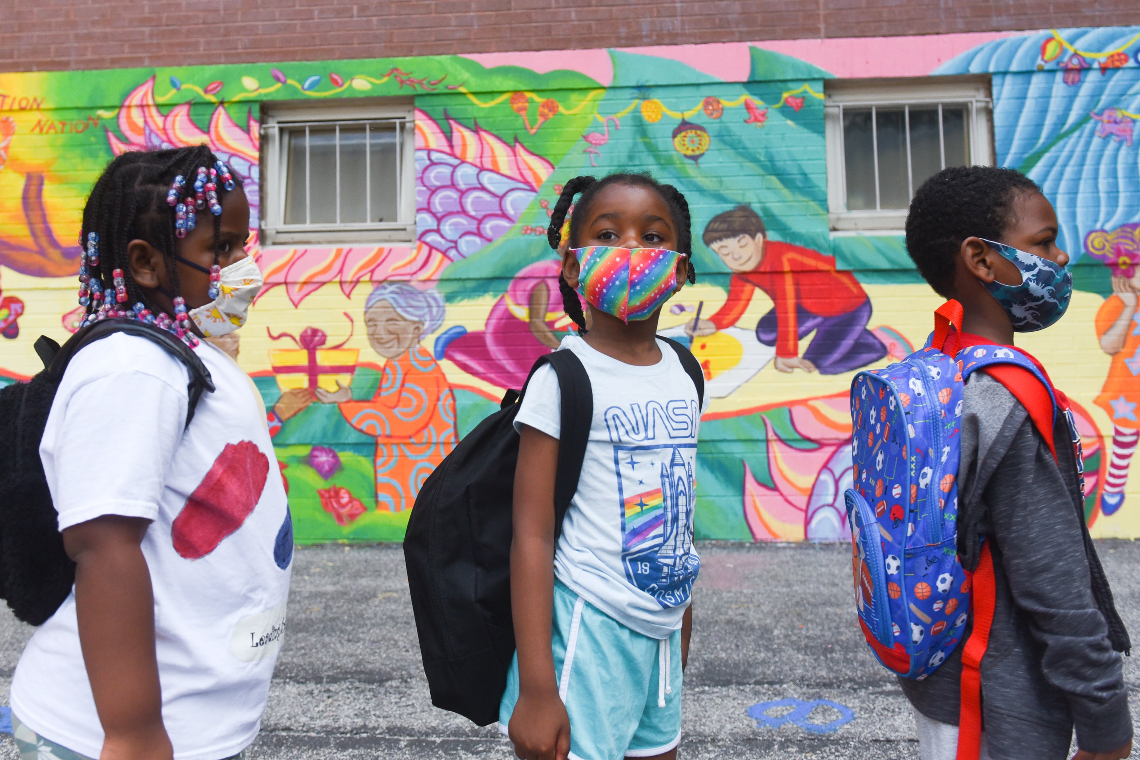 Three masked children stand in front of a brightly colored mural outside.
