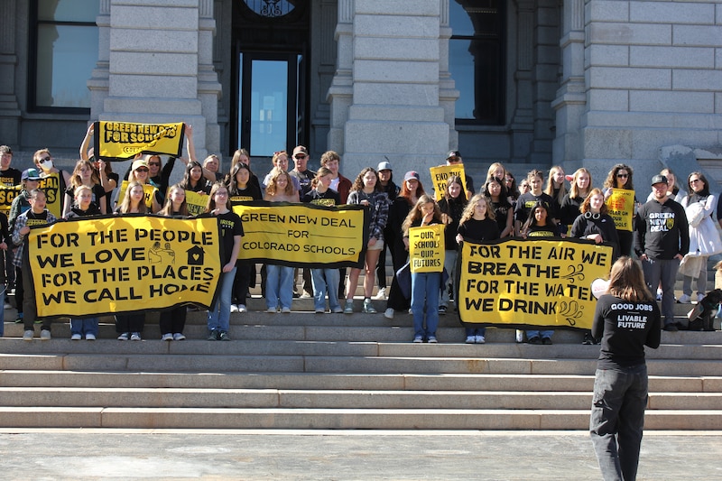 A photograph of a group of students protesting with large yellow signs standing on the steps of the Colorado Capitol building.