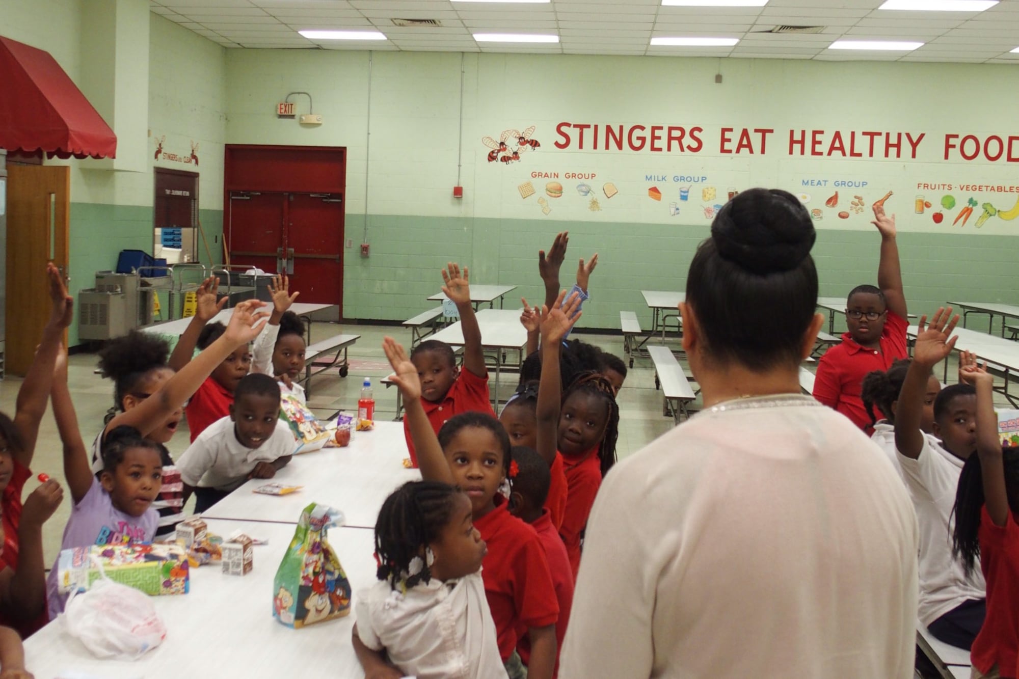Students sitting around a school cafeteria table raise their hands when their principal asks a question