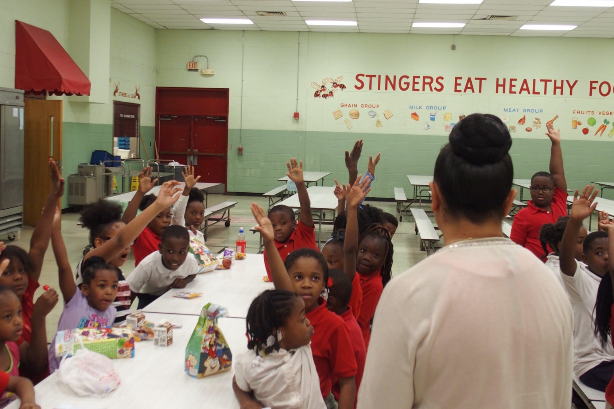 Students sitting around a school cafeteria table raise their hands when their principal asks a question