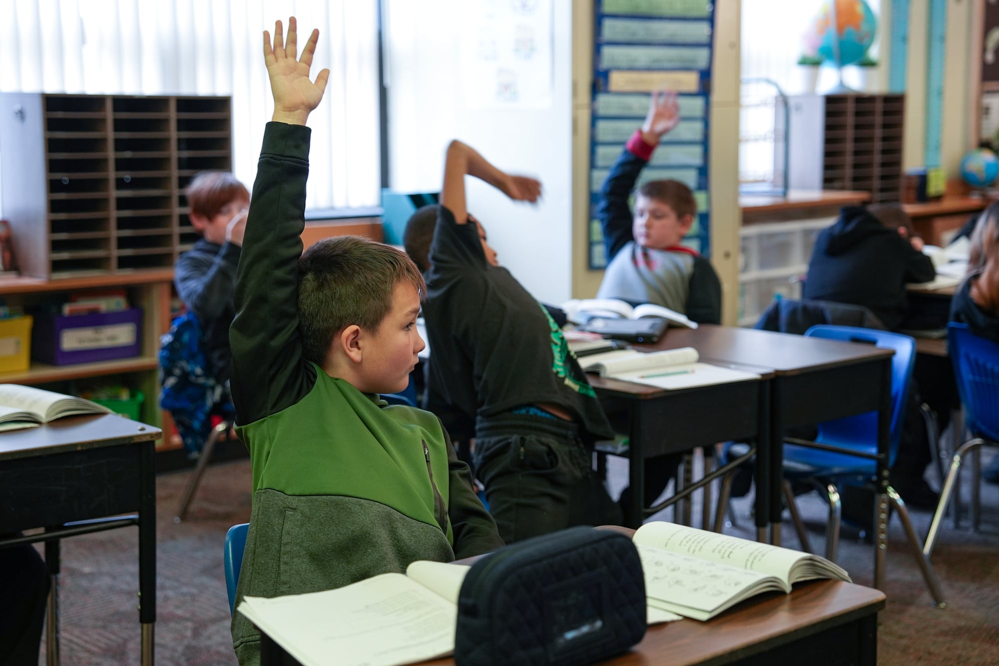 A group of young students sitting at their desks raise their hands in a classroom.
