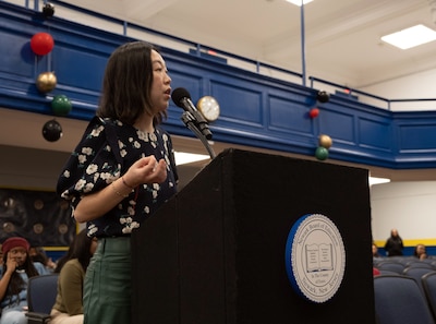 A photograph of a woman speaks from a microphone during a school board meeting in a school auditorium.