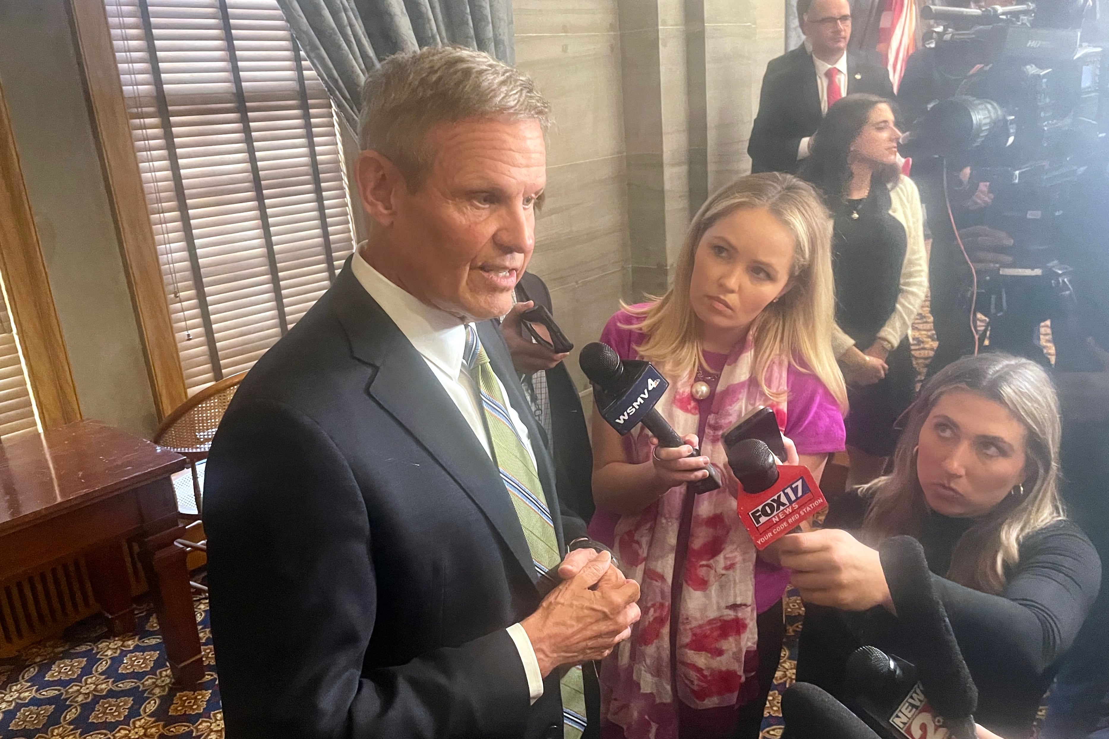 A man wearing a suit is talking to reporters who are holding out microphones.