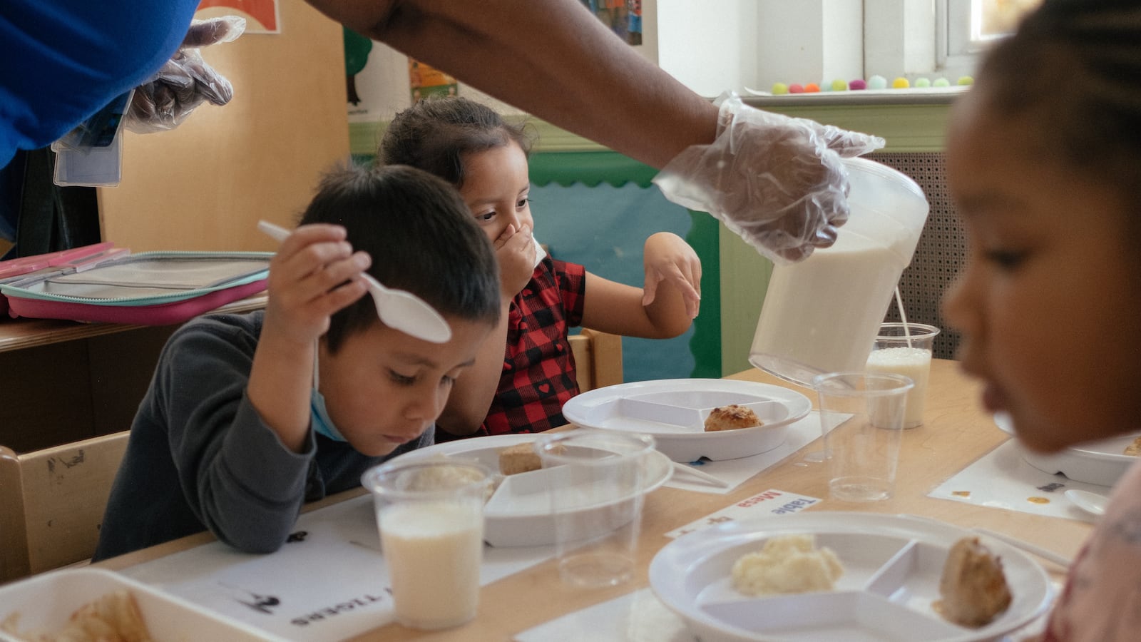 A teacher pours milk into a young student’s cup as three children eat a meal at school.