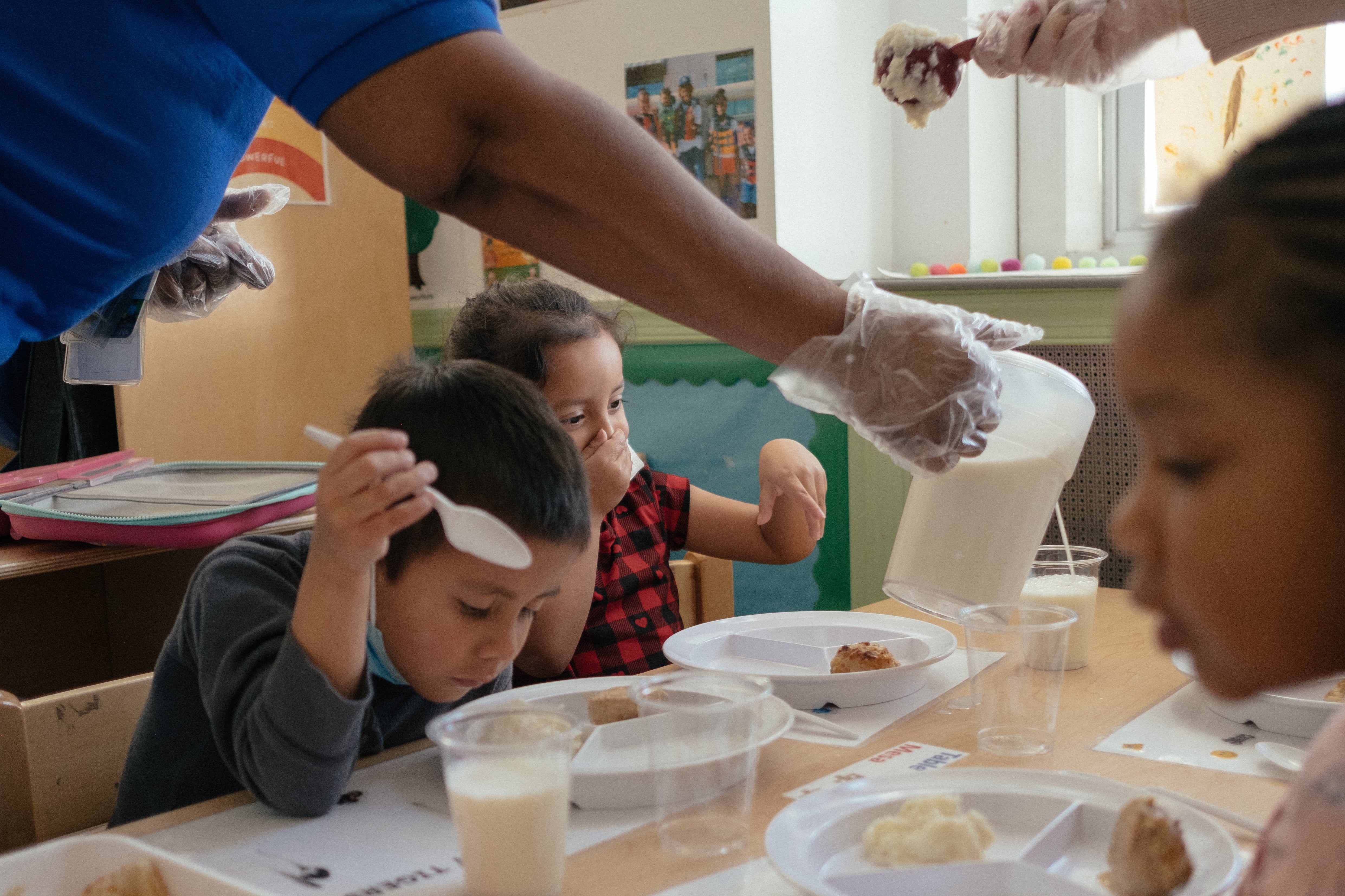 A teacher pours milk into a young student’s cup as three children eat a meal at school.