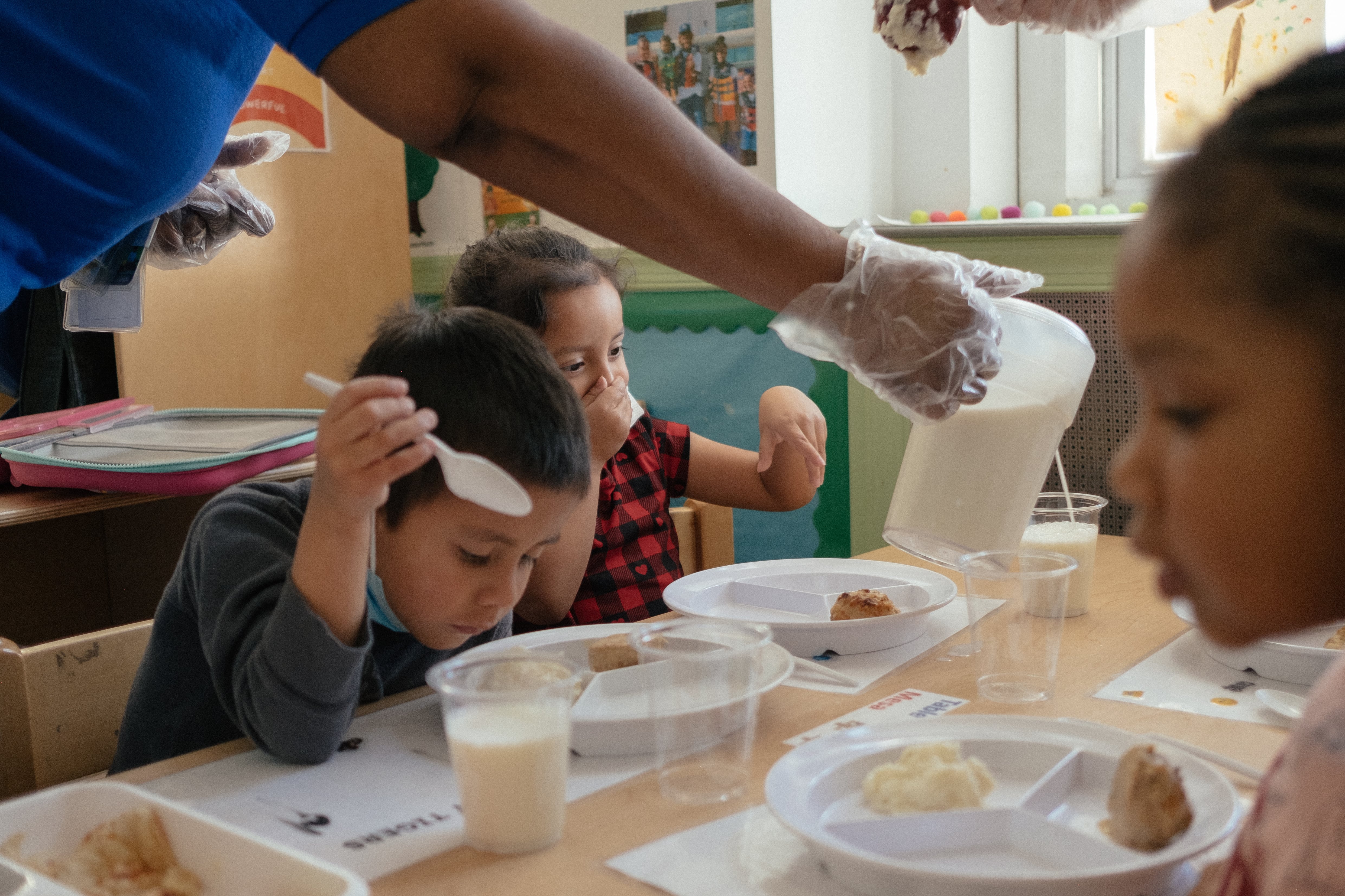 A teacher pours milk into a young student’s cup as three children eat a meal at school.