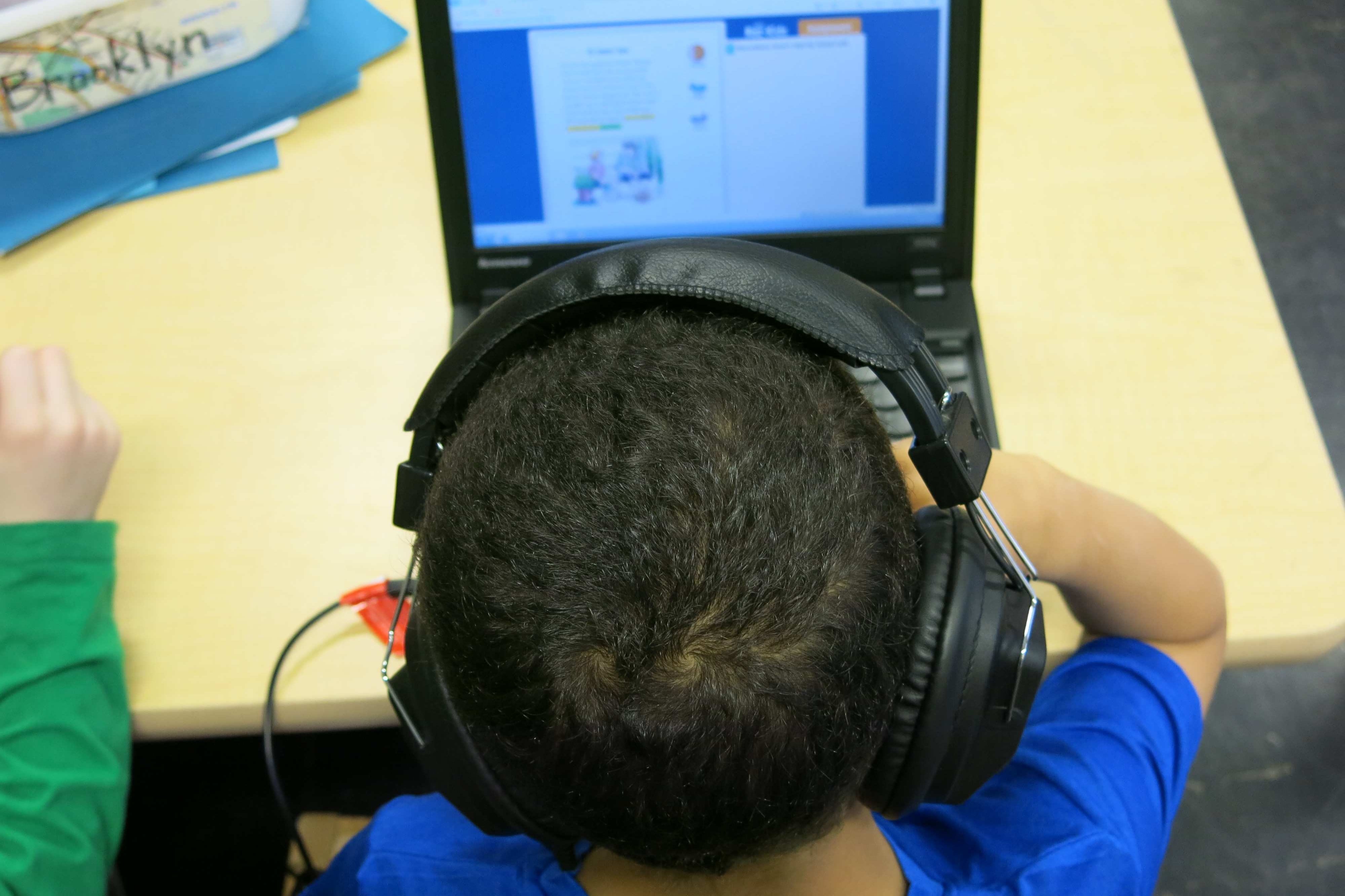 A young student wearing headphones and working on a laptop. The foreground is a close up of the child’s head with the laptop in the background.