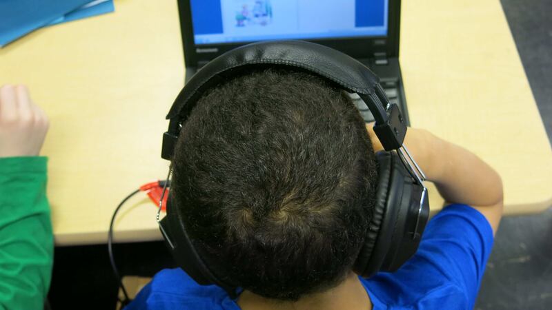 A young student wearing headphones and working on a laptop. The foreground is a close up of the child’s head with the laptop in the background.