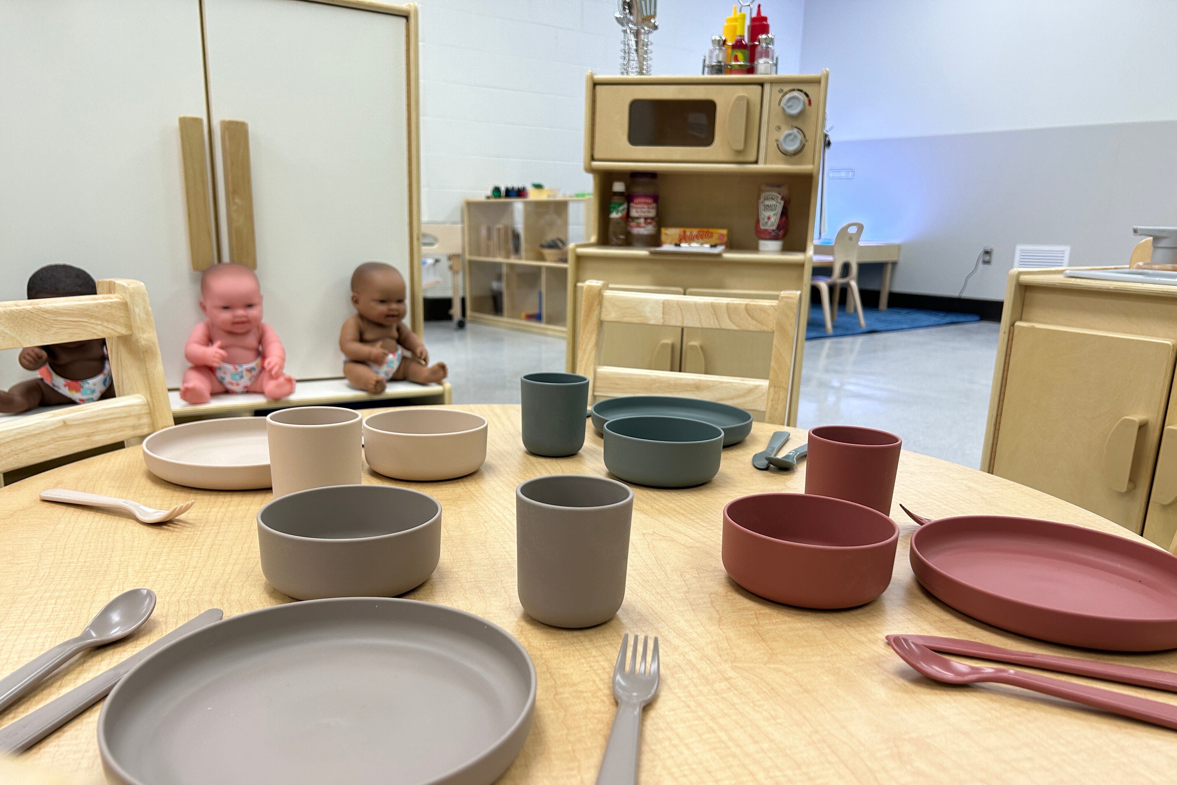 Silverware and plates sit on a children’s dining table in the foreground, with children’s kitchen playware in the background.