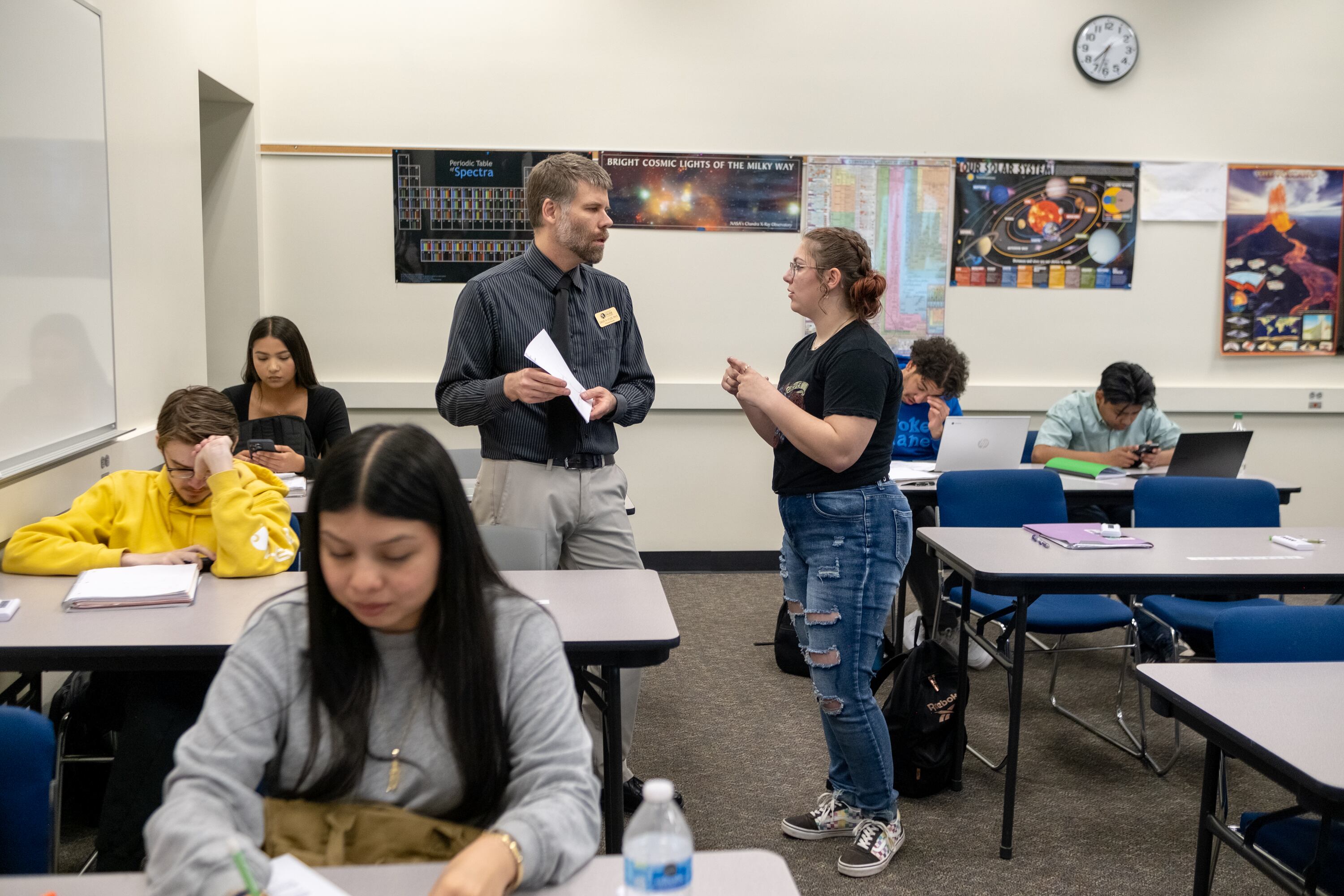 A woman in a black t-shirt and jeans stands in a classroom with a man in a gray shirt and light-colored pants.