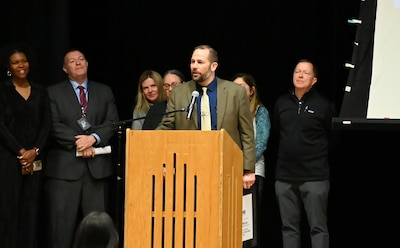 An adult man wearing a suit stands behind a wooden podium with a group of people standing behind him.