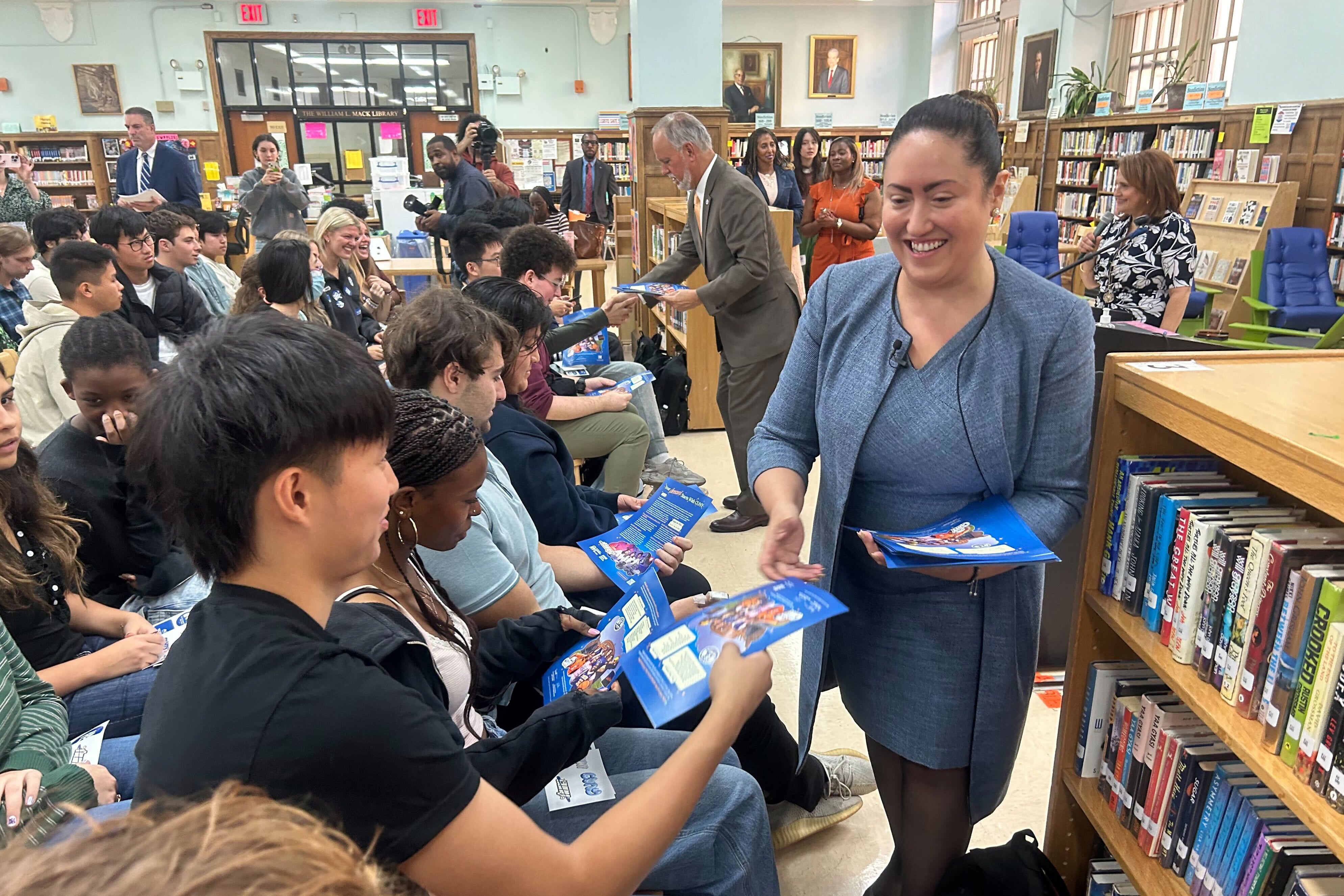 A woman wearing a blue dress suit hands out pieces of paper to students who are all sitting down in a school library.