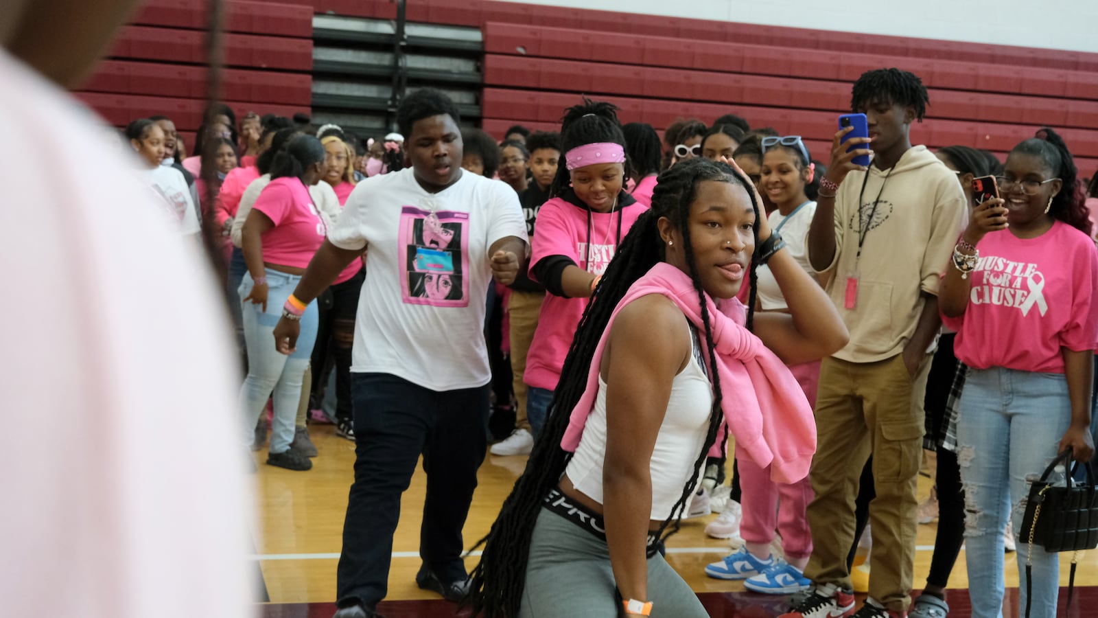 A student wearing a pink sweater around their neck dances in the middle of a circle of students who are filming on their phones and also dancing.