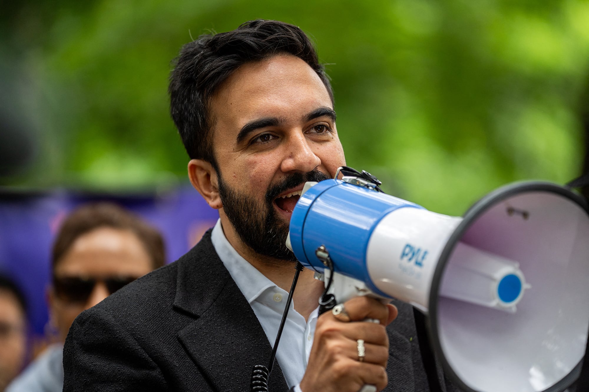 A man wearing a suit speaks into a megaphone outside with green trees in the background.