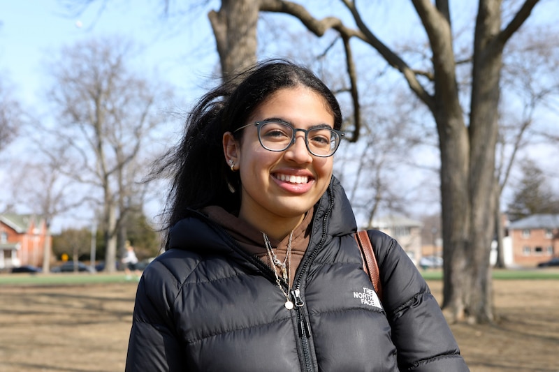 A high school student with dark hair tied up and wearing a black jacket smiles for a portrait outside on a cold and sunny day.