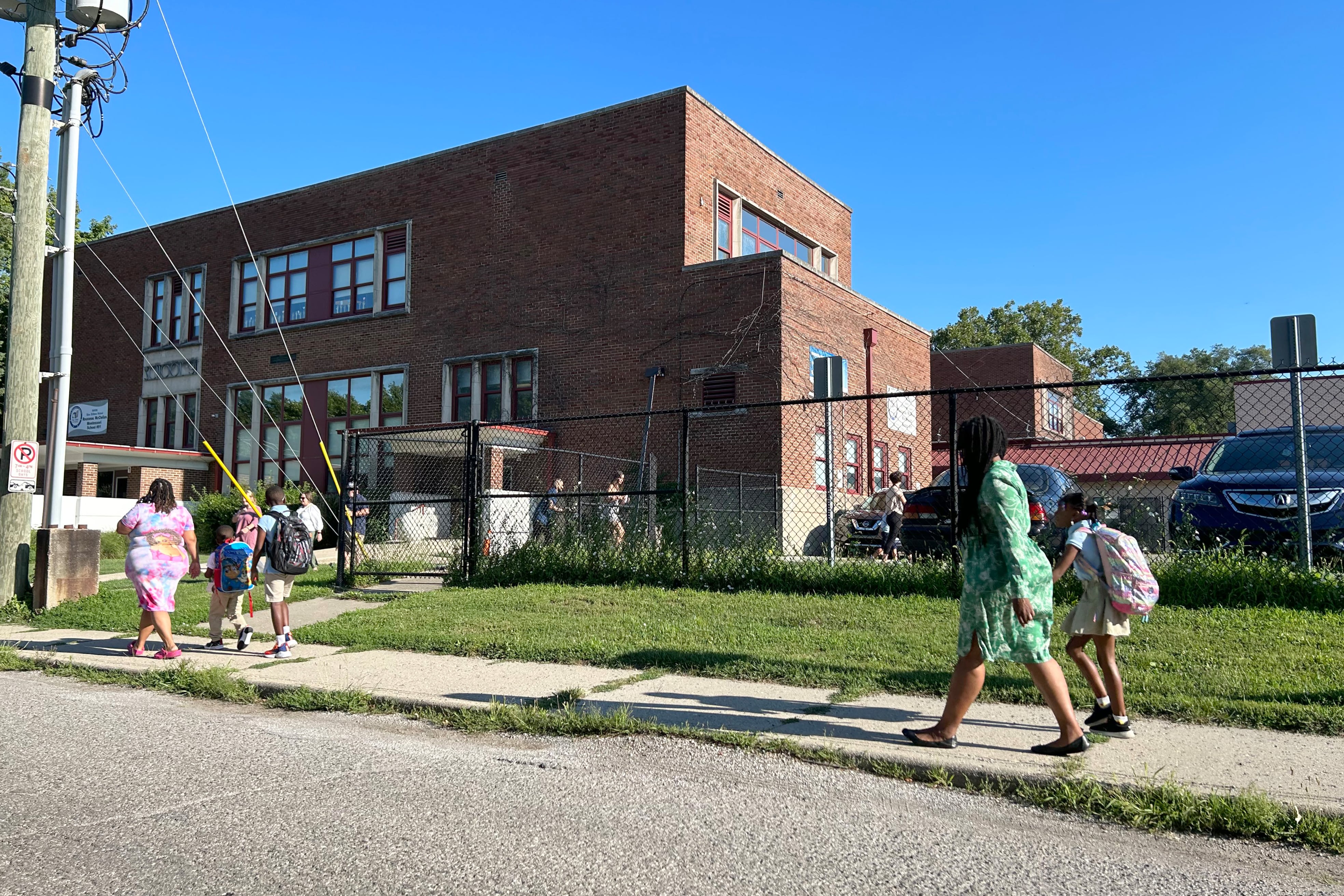 Five people walk on a sidewalk heading into a brick school building.