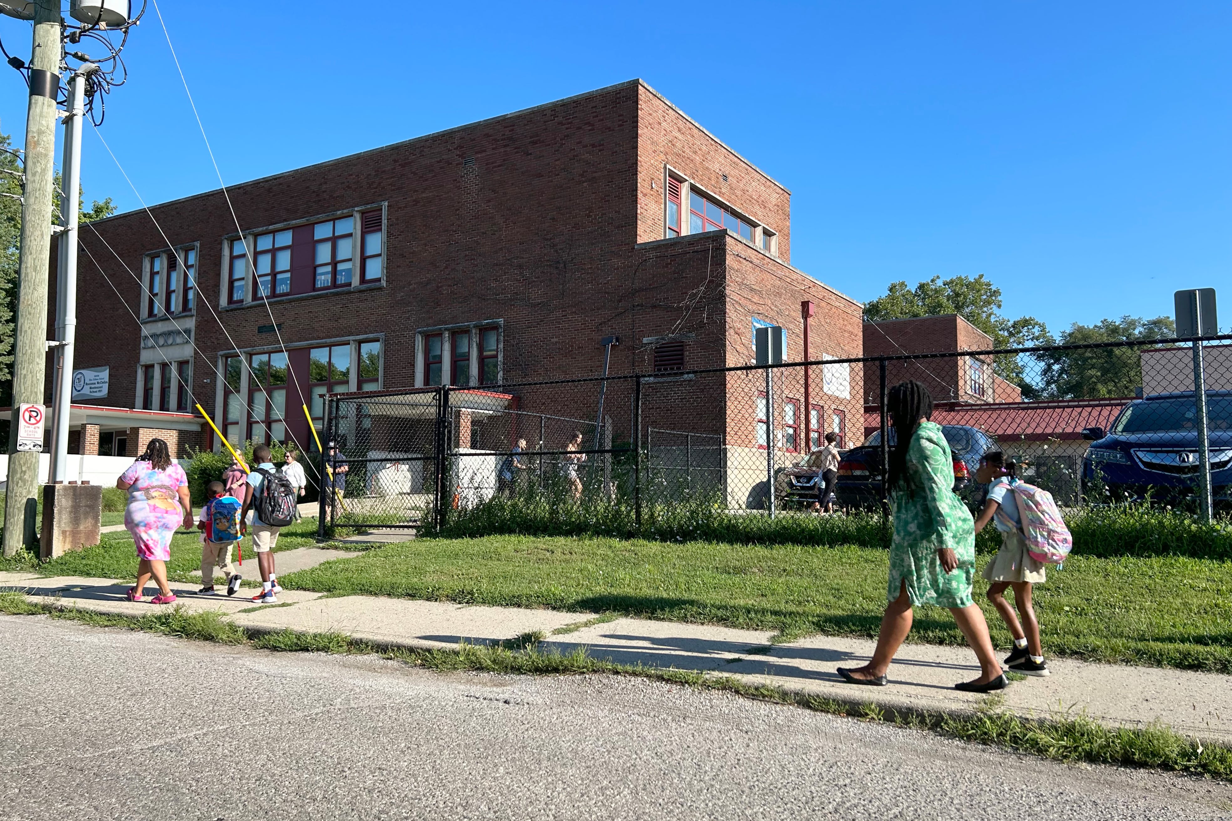 Five people walk on a sidewalk heading into a brick school building.