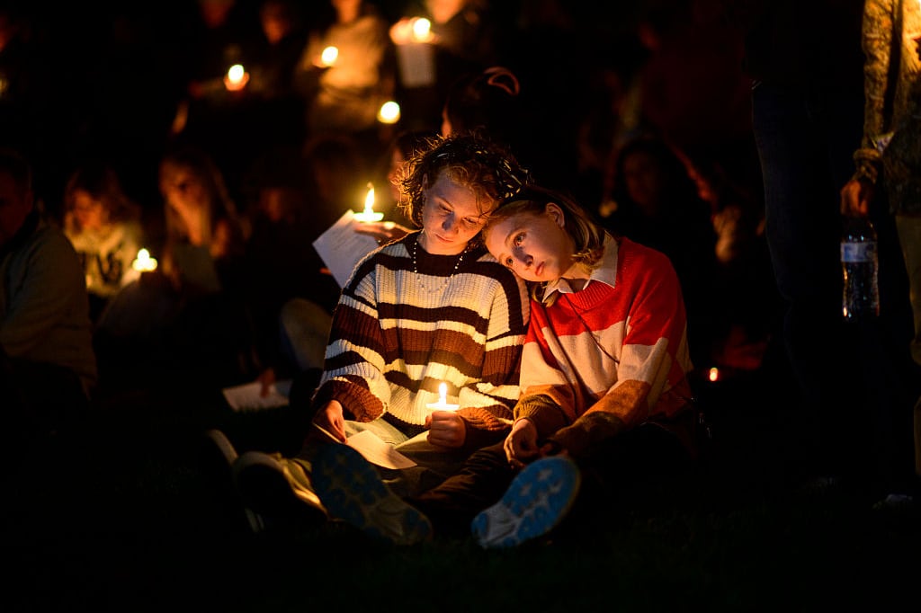 Two girls comfort each other while holding a candle at a nighttime vigil. The flame lights their faces in the darkness. The lights of other candles are visible around them.