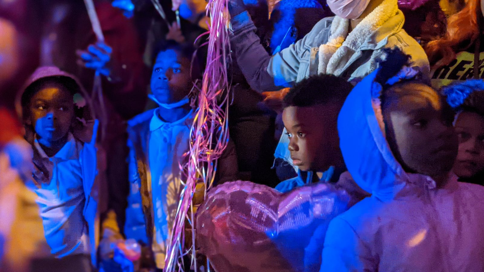 The little brother of shooting victim Phillexus Buchanan and young children stand in front of loved ones during a vigil, with blue light shining over them, and the strings of pink balloons hanging from grasped hands.
