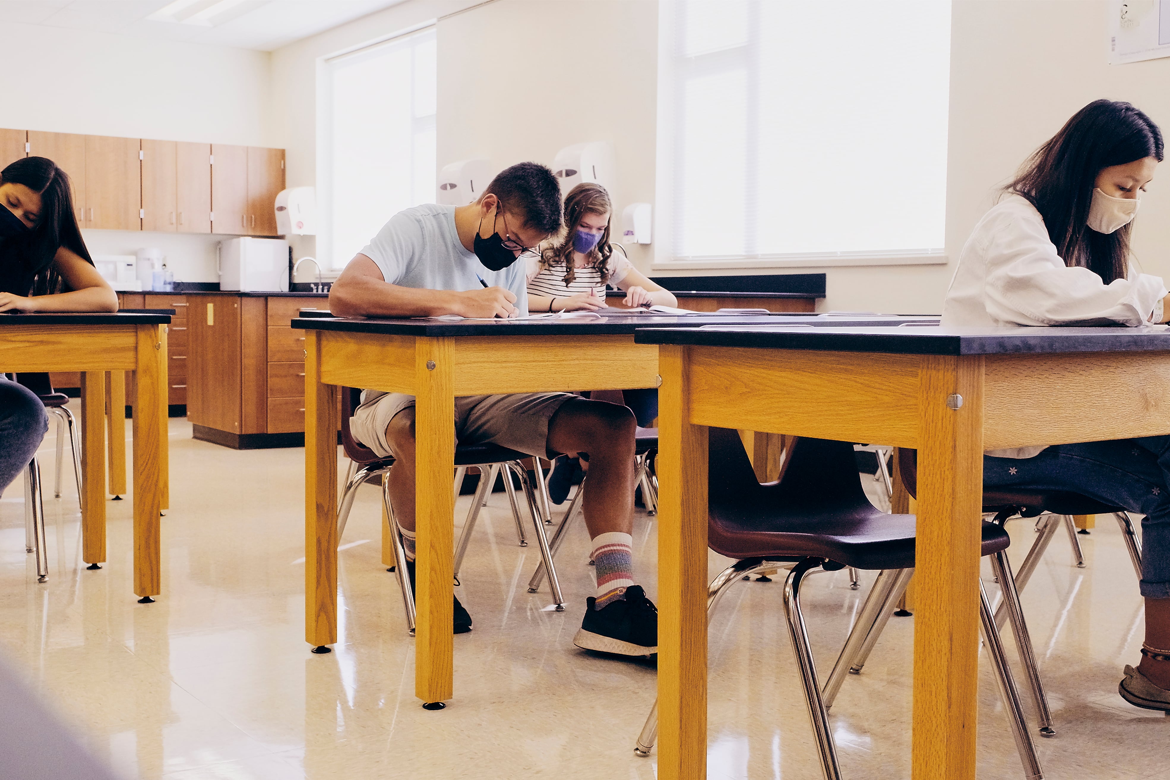 Four masked students write at desks in a classroom.