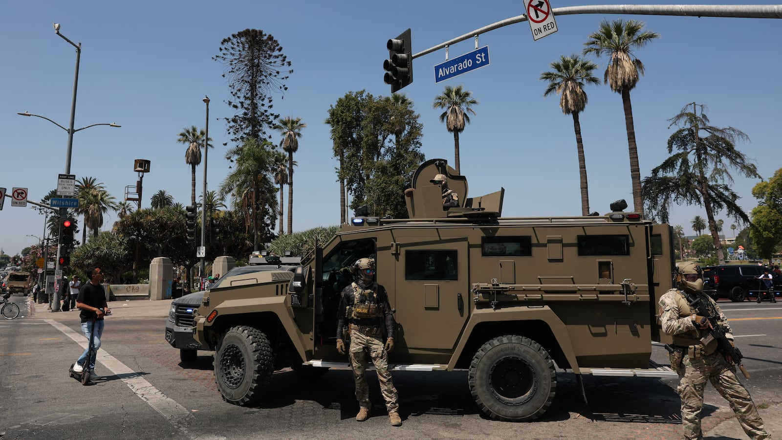 Federal agents with their faces covered stand before an armored vehicle. There are palm trees in the background. A street sign reads Alvarado Street.