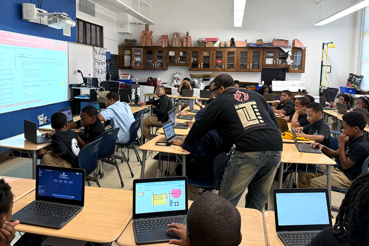 A man in a black t-shirt works with a student at a desk in a classroom.