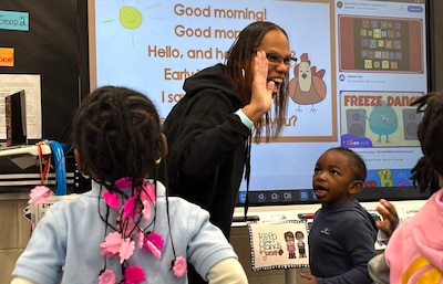 A woman with a dark shirt smiles at her students in a classroom.