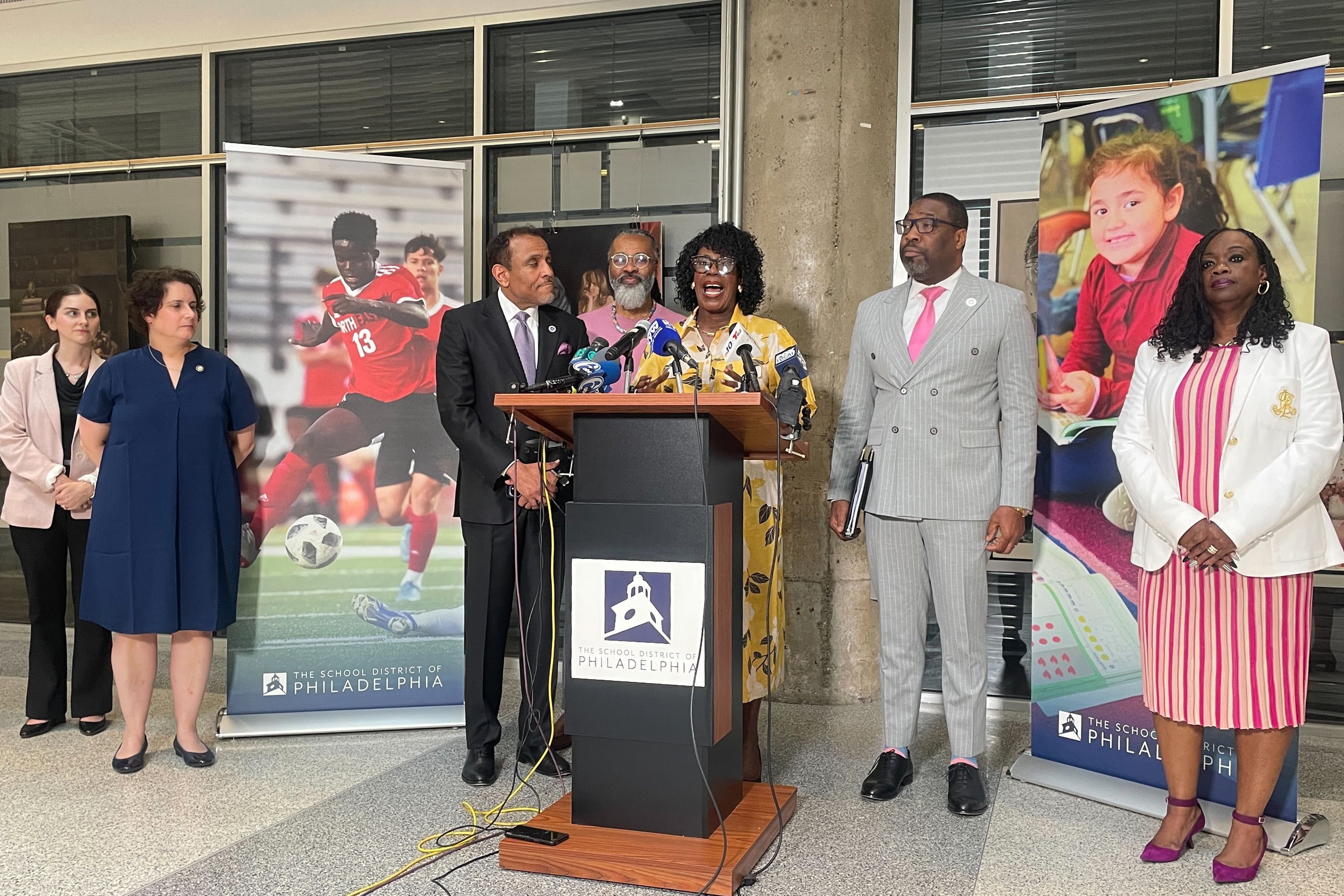 A photograph of a group of adults in business clothes while one Black woman in a yellow dress speaks from behind a wooden podium in front of a building.