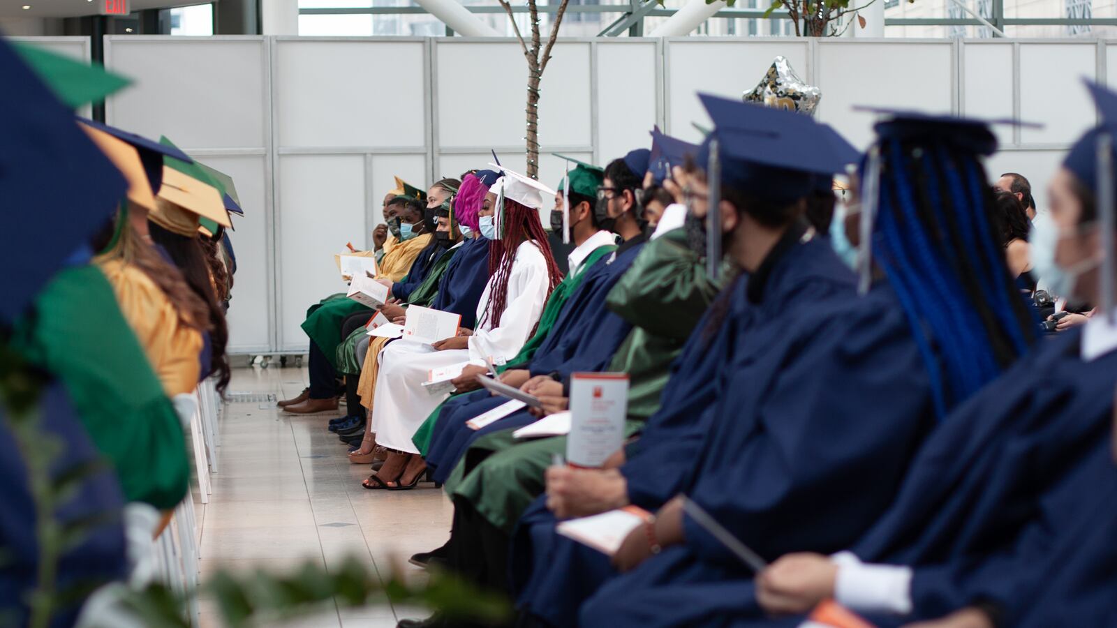 Rows of graduating students wearing blue, yellow, white and green graduation gowns and caps listen to a speaker.