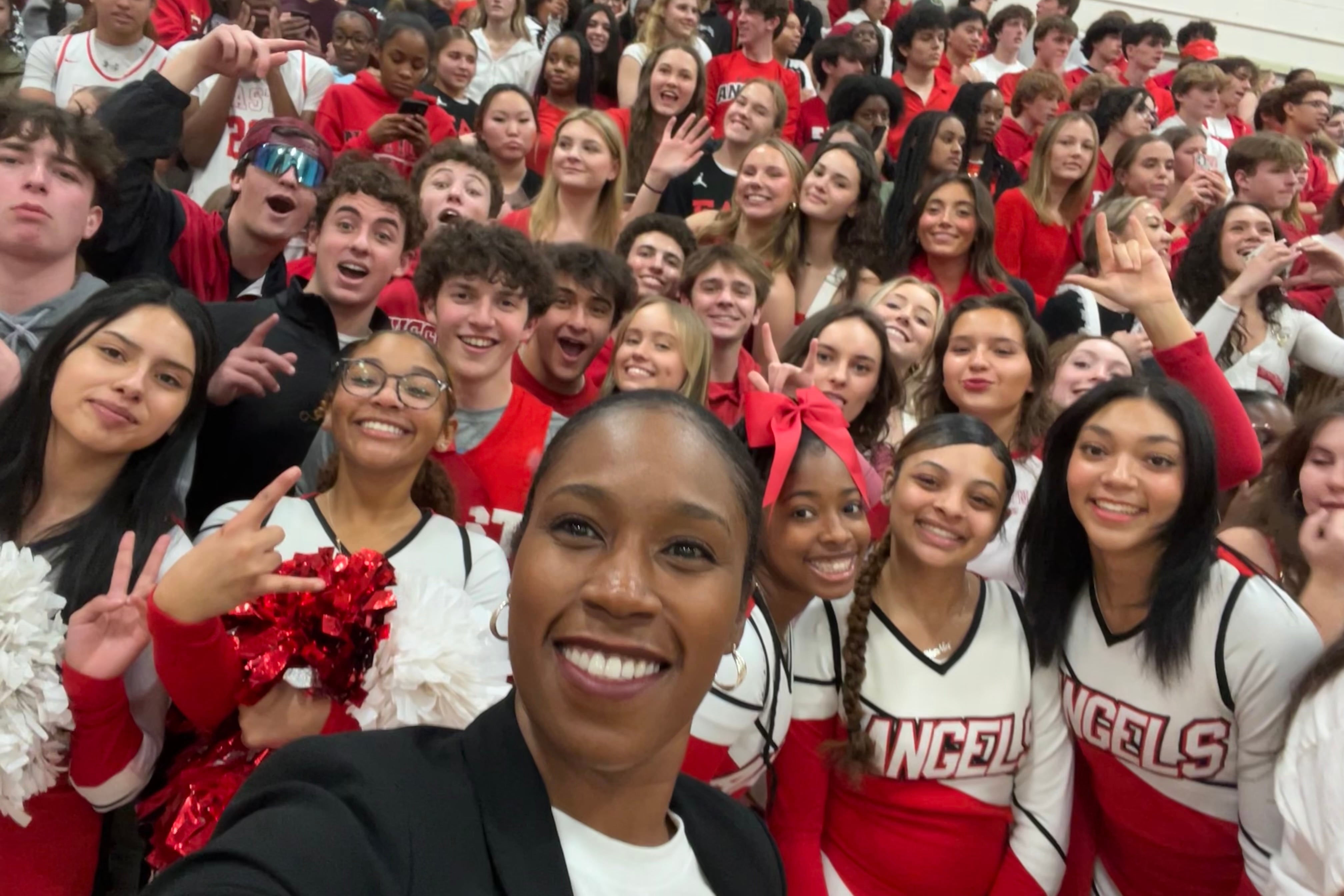 A Black woman takes a selfie with a large group of high school students all wearing red and inside of a gym.