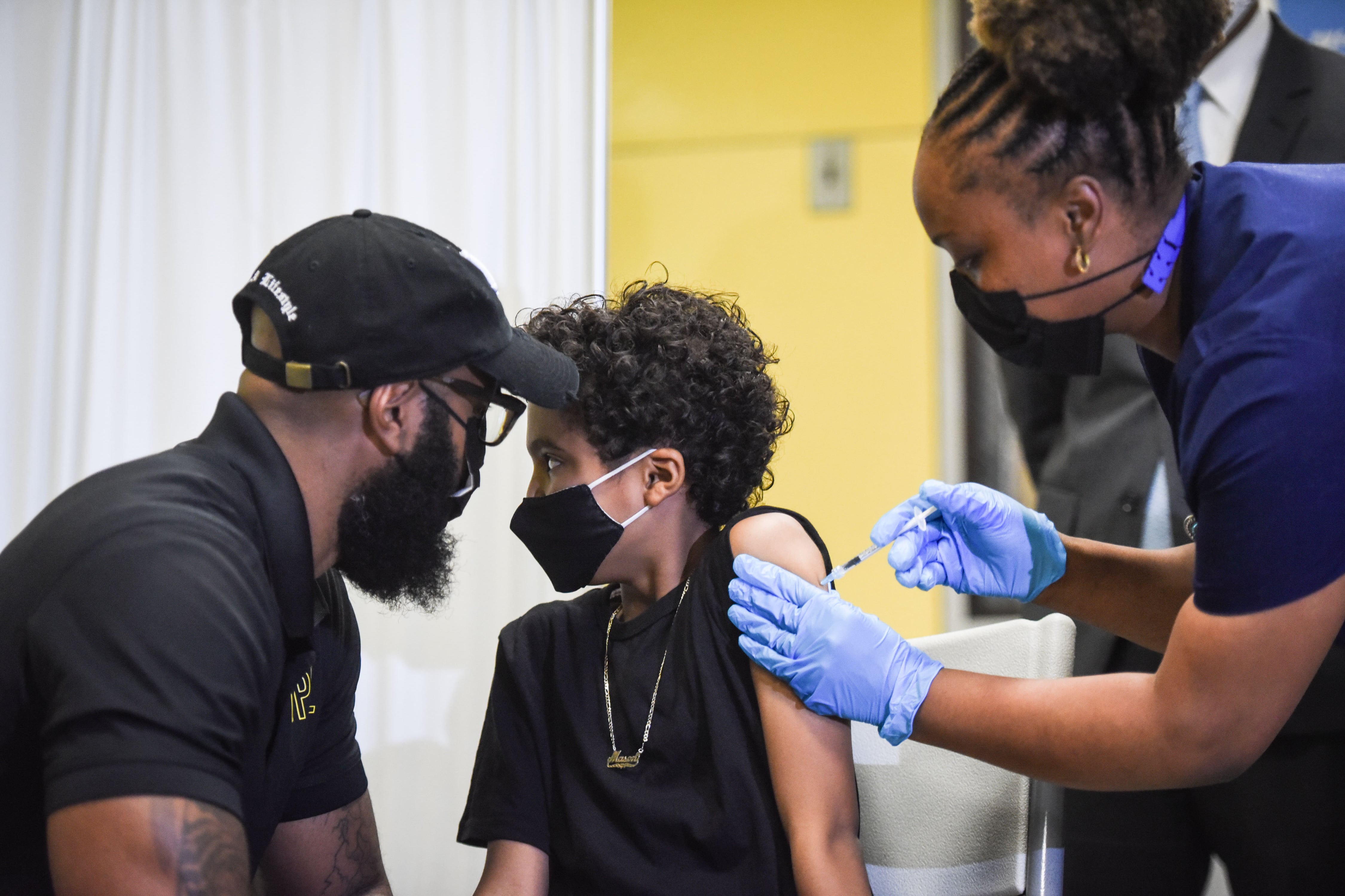 A boy receives a dose of the COVID vaccine from a health care professional as a man looks on next to him.