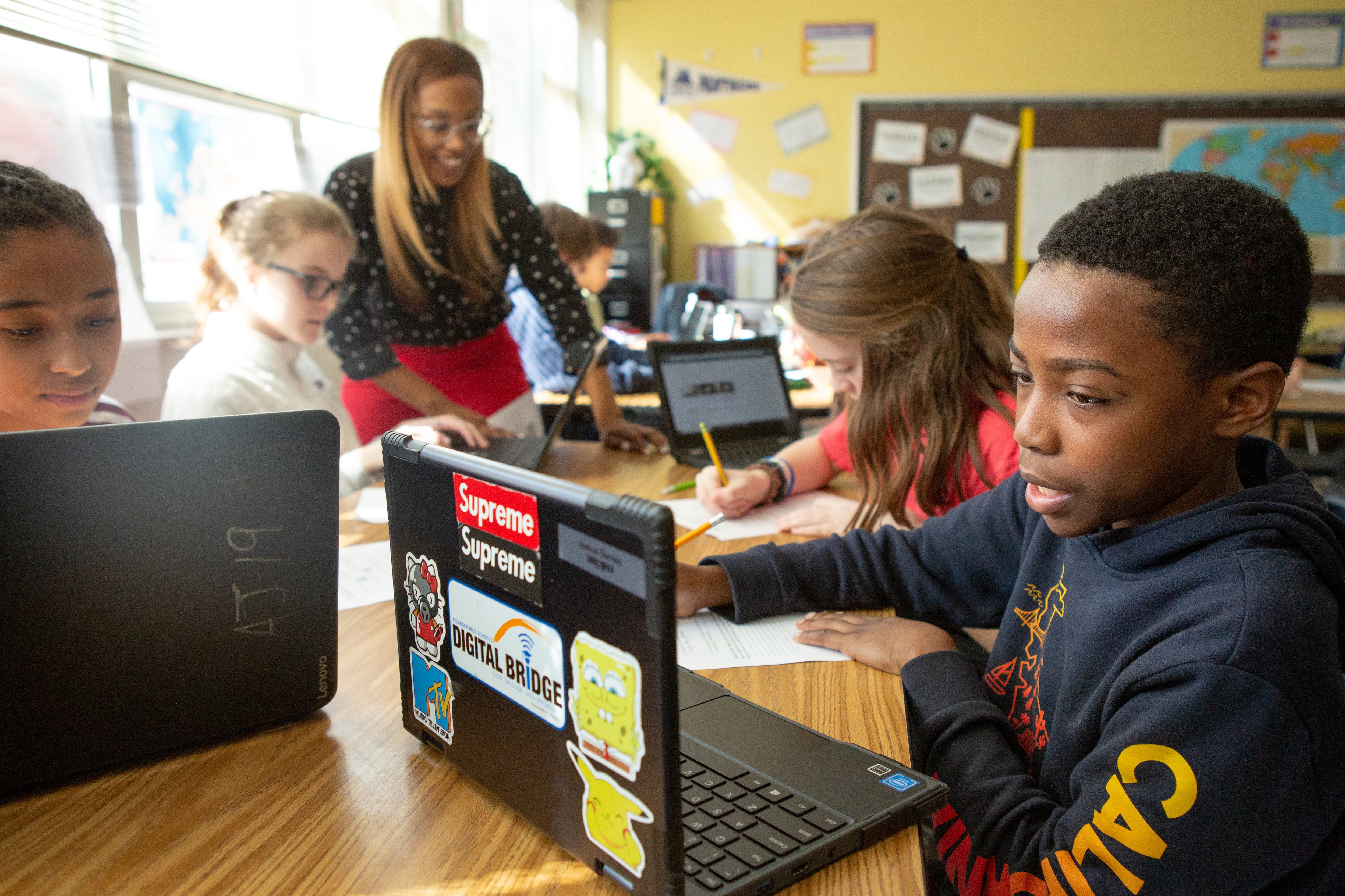 Students sit at a table with laptops with an adult helping a student in the background.