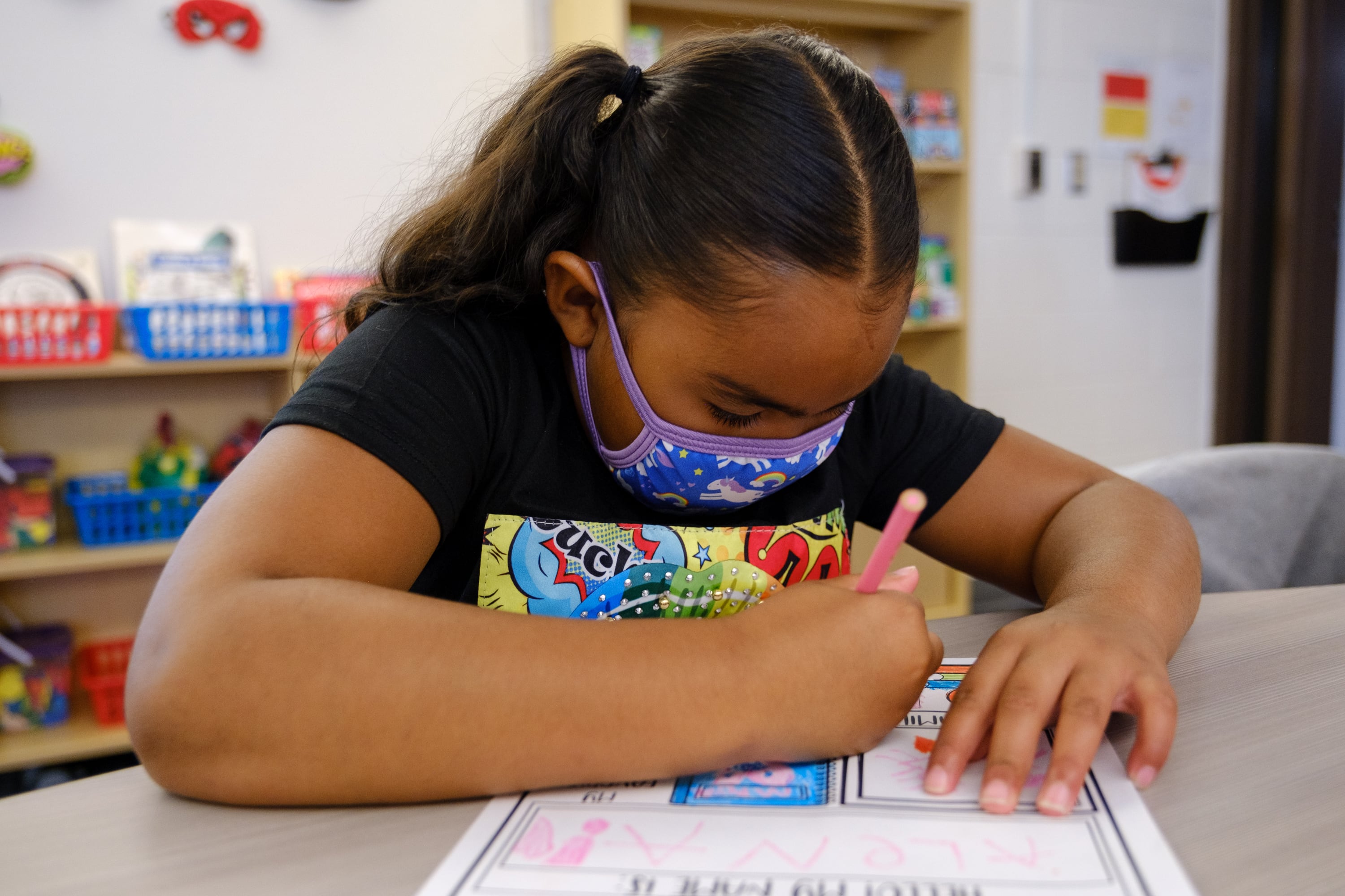 A young girl wearing a mask writes on a worksheet at her desk.
