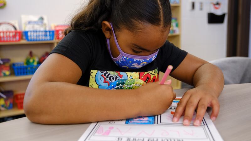 A young girl wearing a mask writes on a worksheet at her desk.