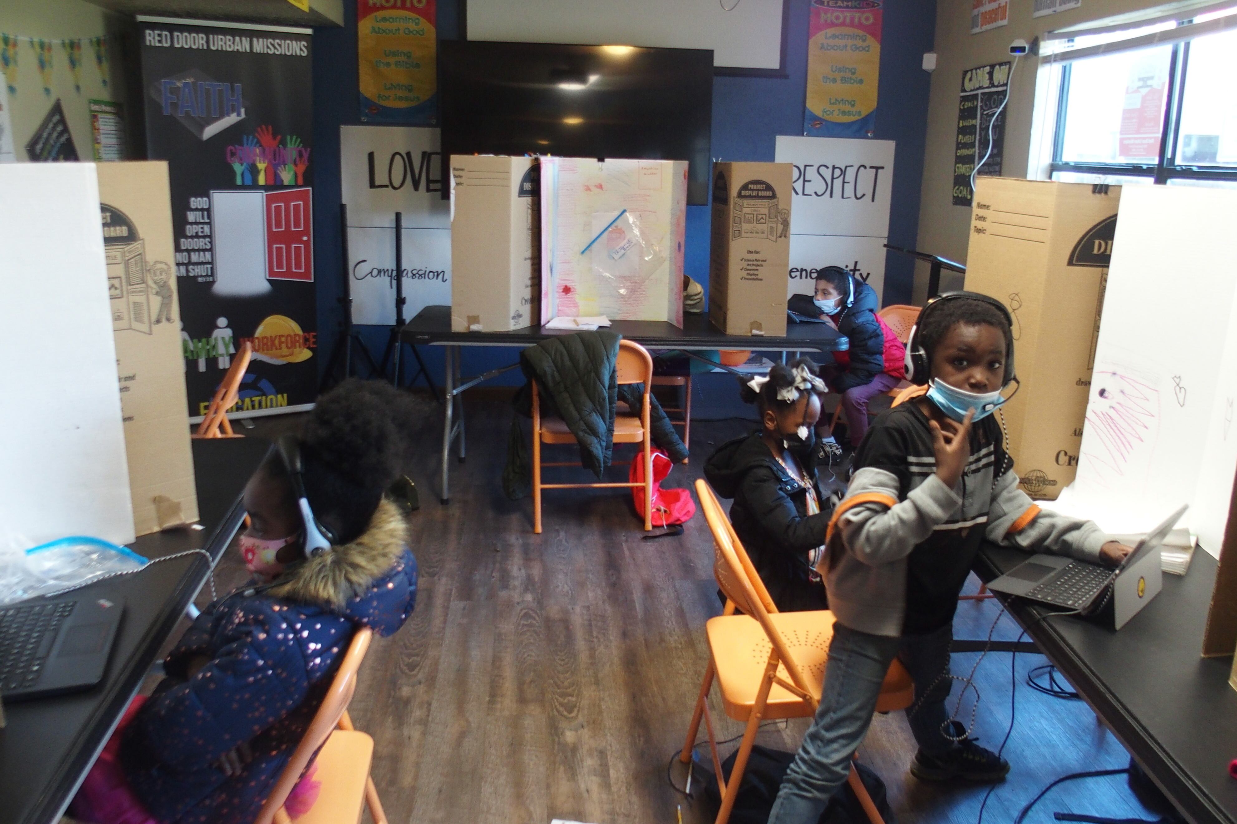 Four children wearing masks and headphones work at laptop computers in a narrow classroom with orange folding chairs. Three hand-lettered posters on the walls read “Respect,” “Love,” and “Compassion.” 