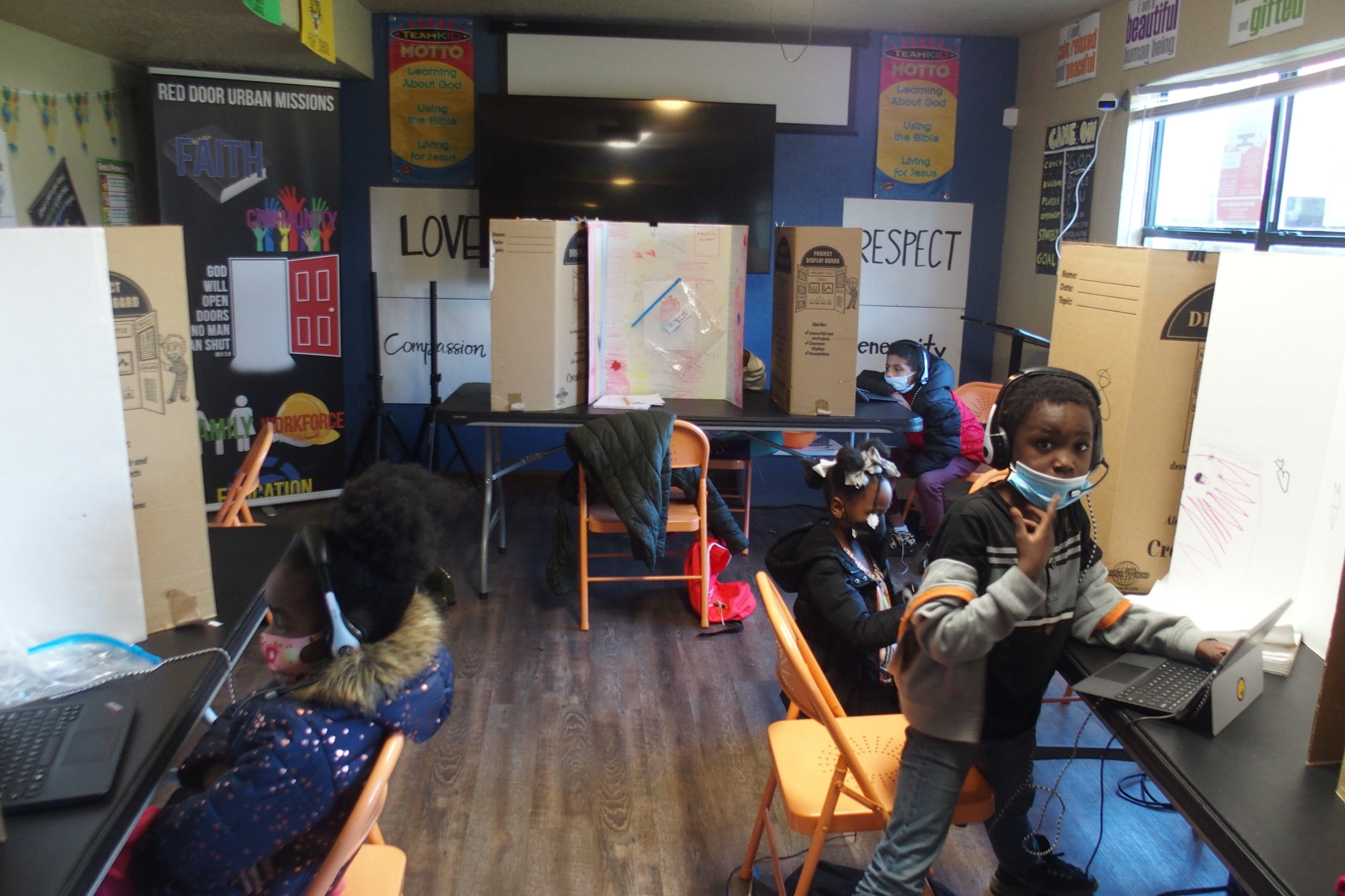 Four children wearing masks and headphones work at laptop computers in a narrow classroom with orange folding chairs. Three hand-lettered posters on the walls read “Respect,” “Love,” and “Compassion.”