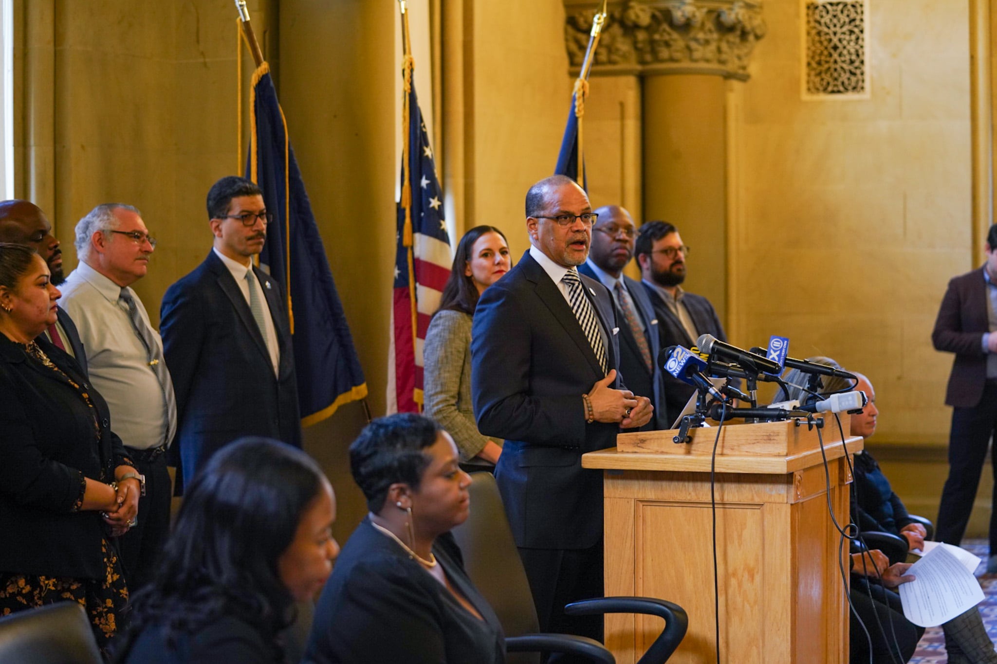 A man wearing a dark suit stands at a podium while many people stand behind him. There is an American flag and two other flags in the background.