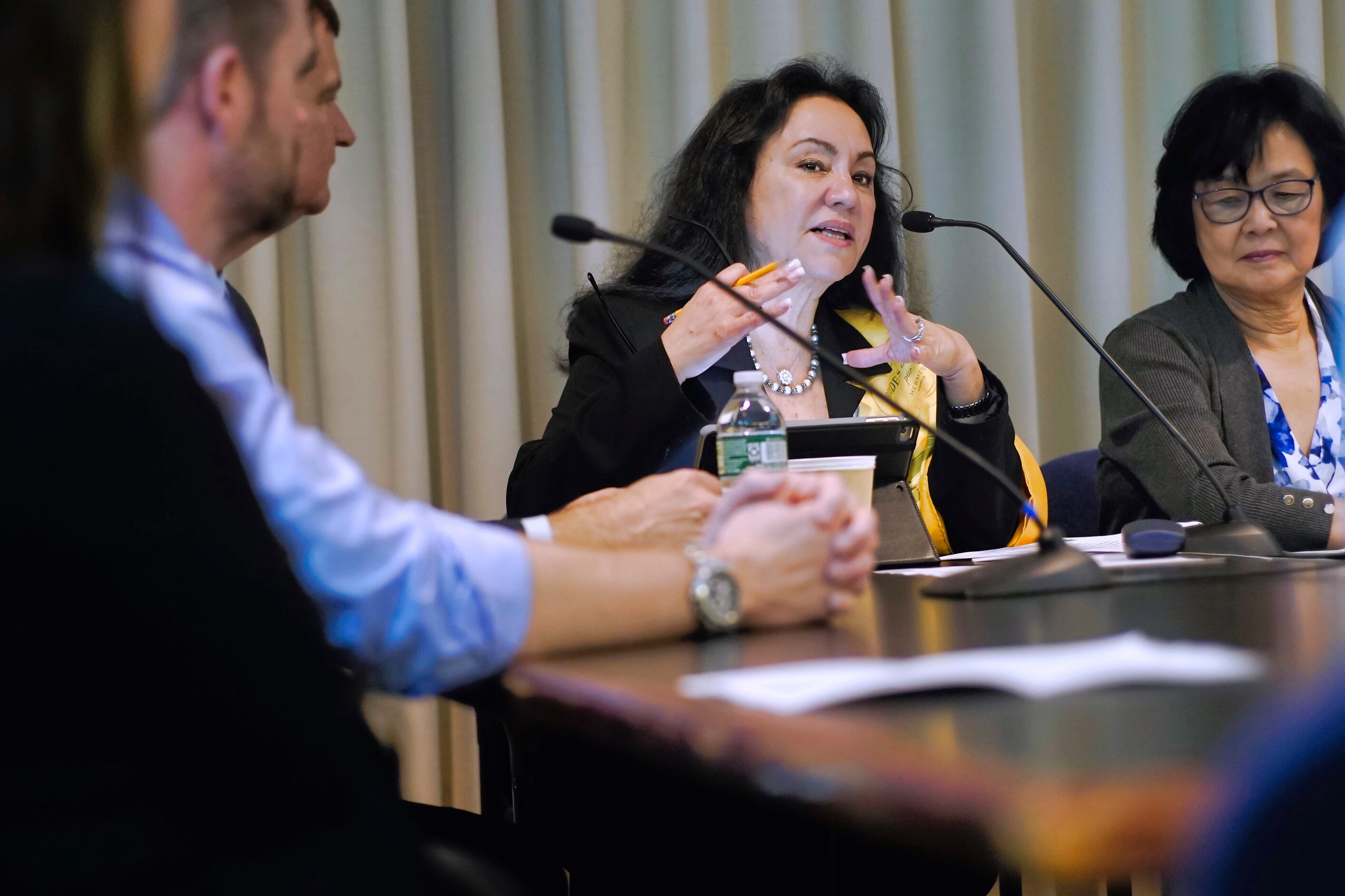 Three people in business attire sit at a wooden desk with two microphones.