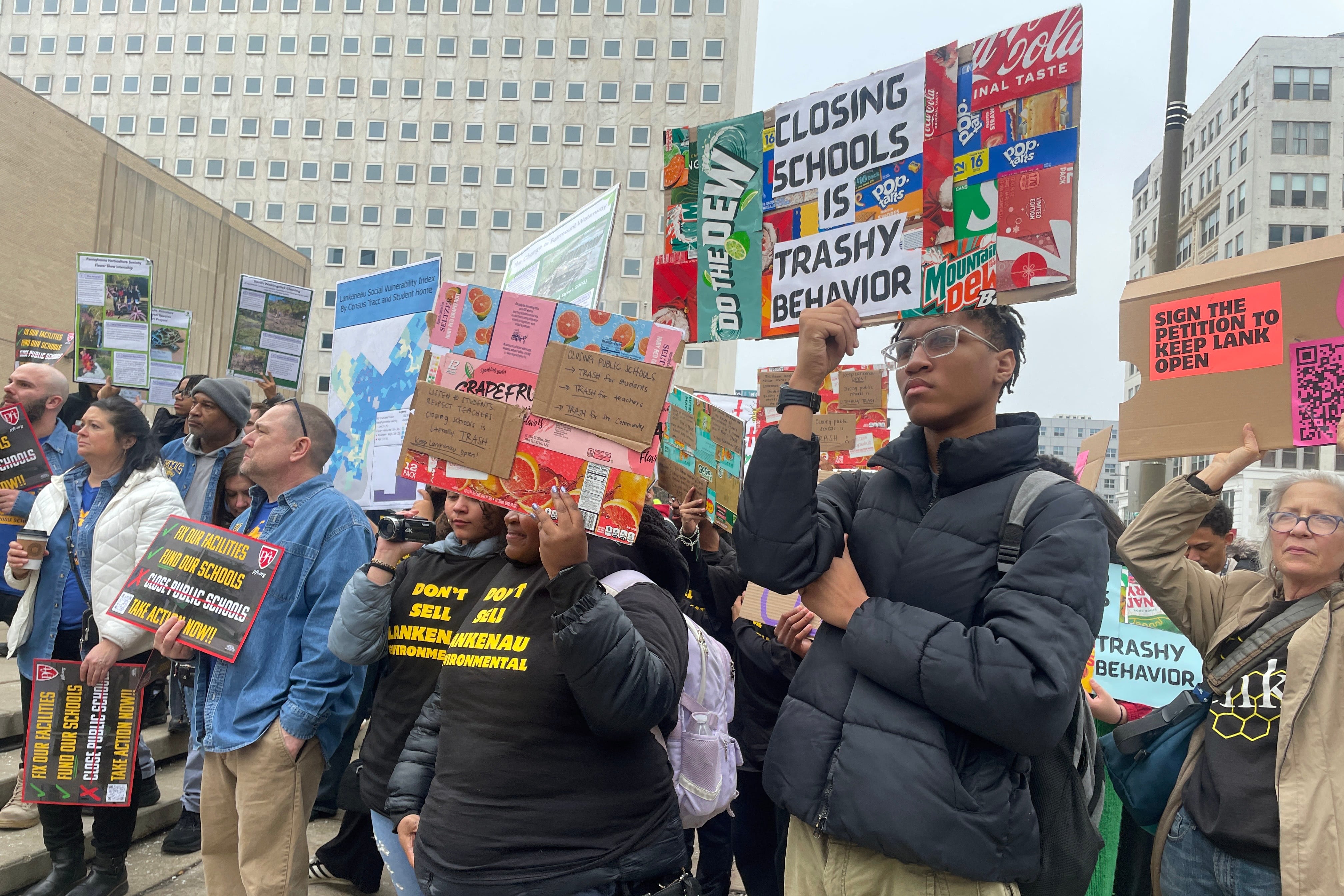 A photograph of a large group of students and adults holding protest signs outside on a cloudy, cold day.