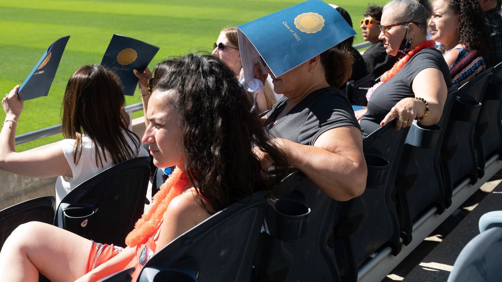 Family members sit in stadium seating and use graduation programs to shade themselves.