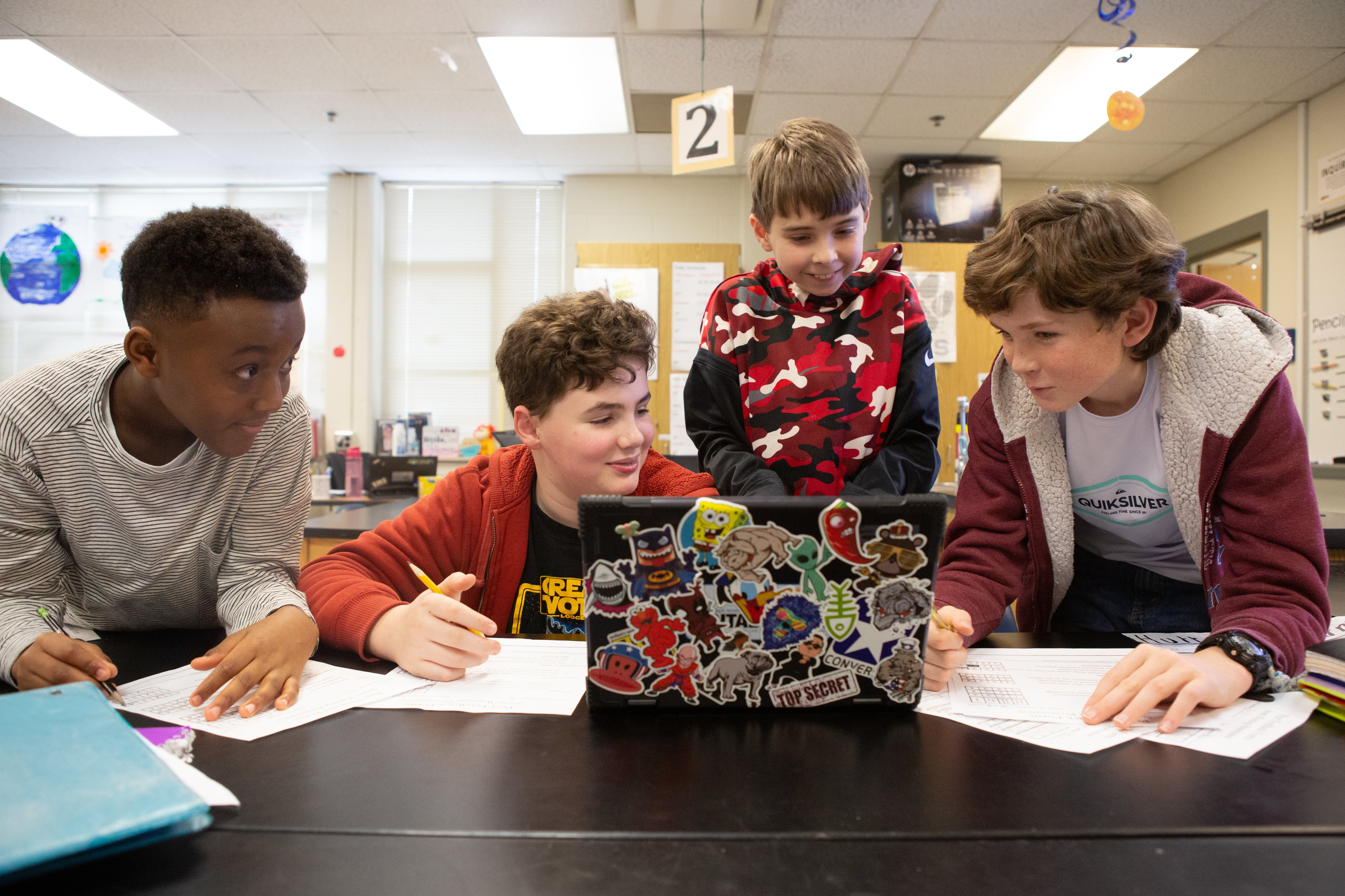 Four middle school students work together on worksheets and a laptop on a desk.