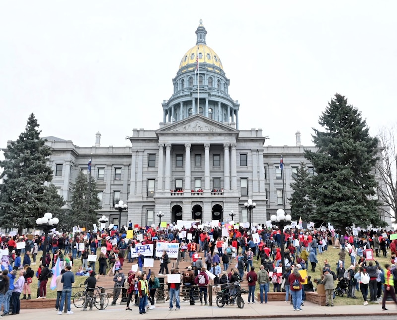 A large group of protestors, some wearing red and some holding signs stand outside of a large Capitol building with a gold dome.