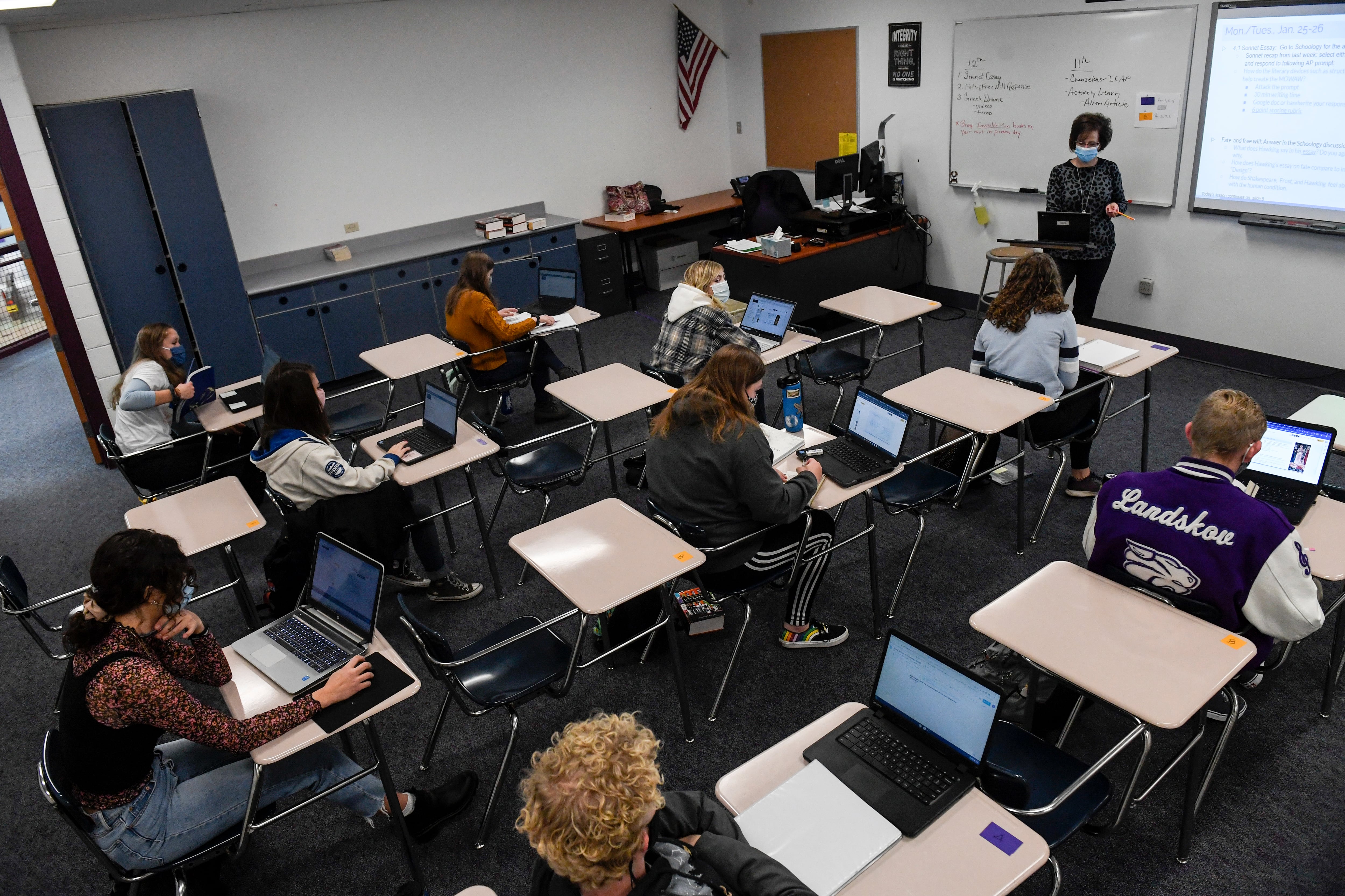 A masked teacher and students use every other desk in a classroom as a COVID precaution.