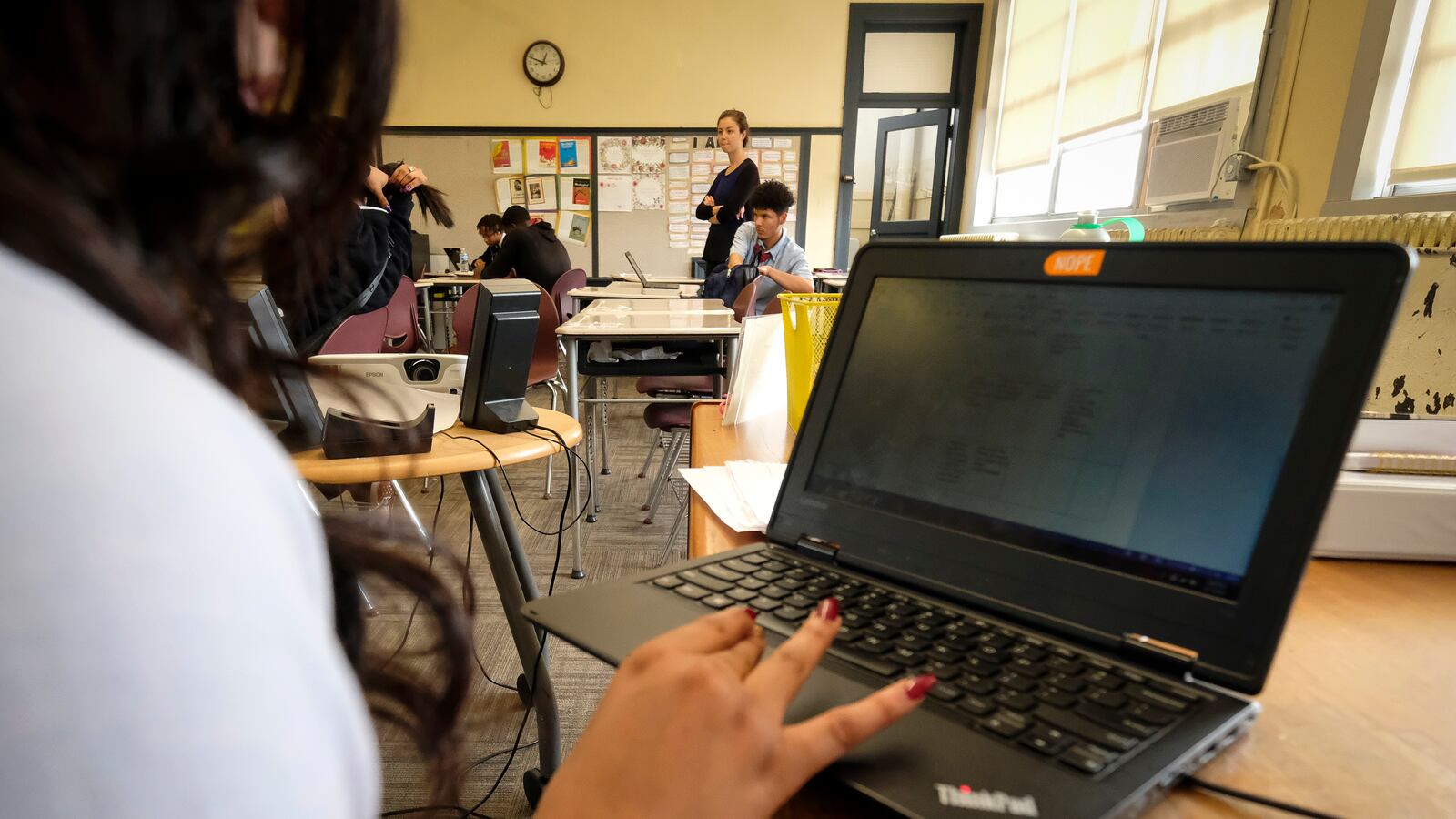 A female student in a classroom uses a laptop computer, with other people in the background.