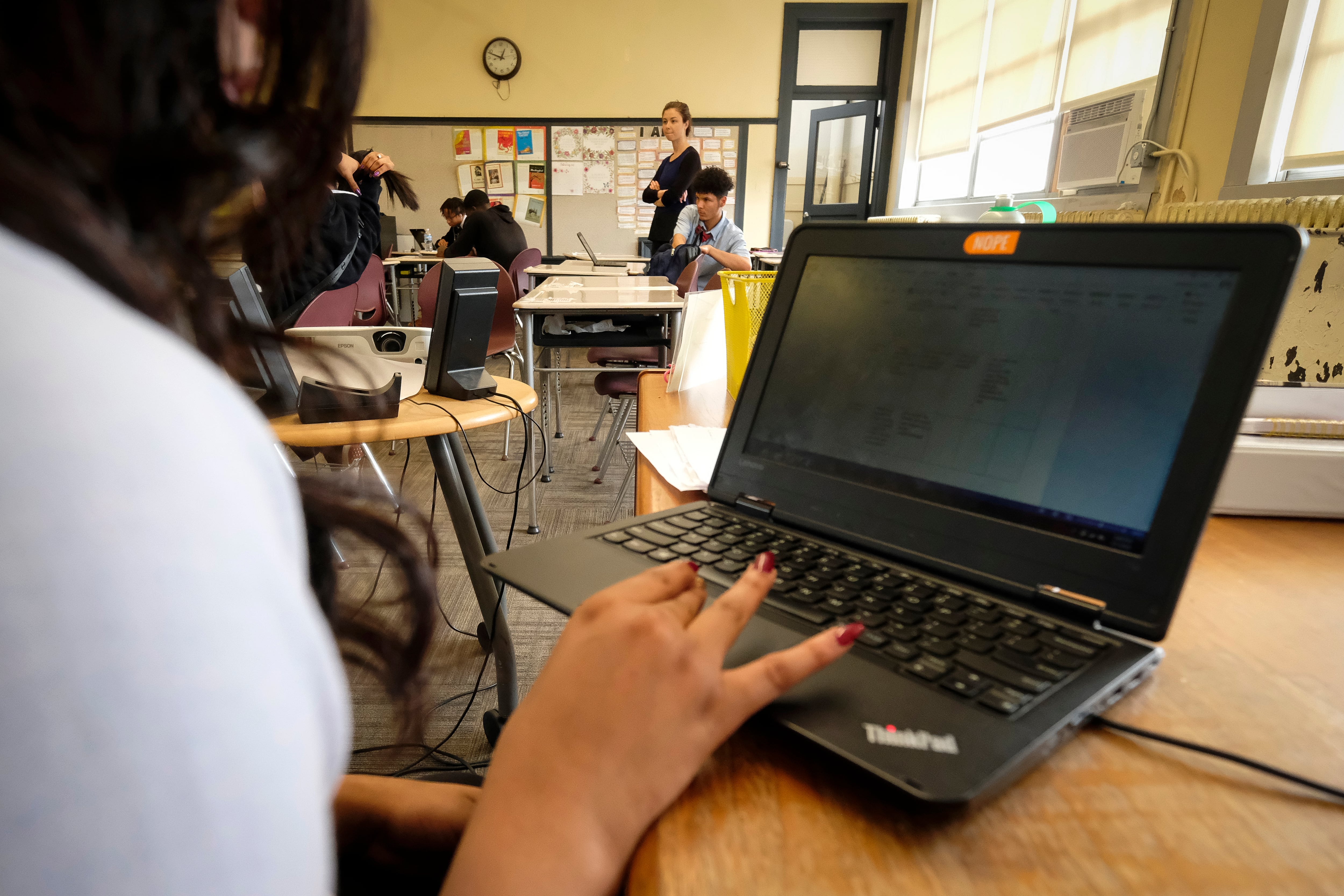 A female student in a classroom uses a laptop computer, with other people in the background.